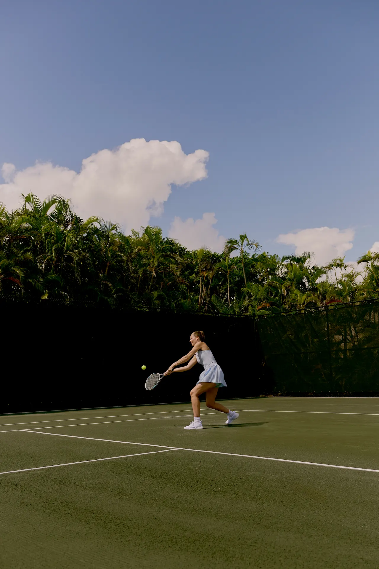Woman playing tennis on outdoor court surrounded by palm trees under blue sky