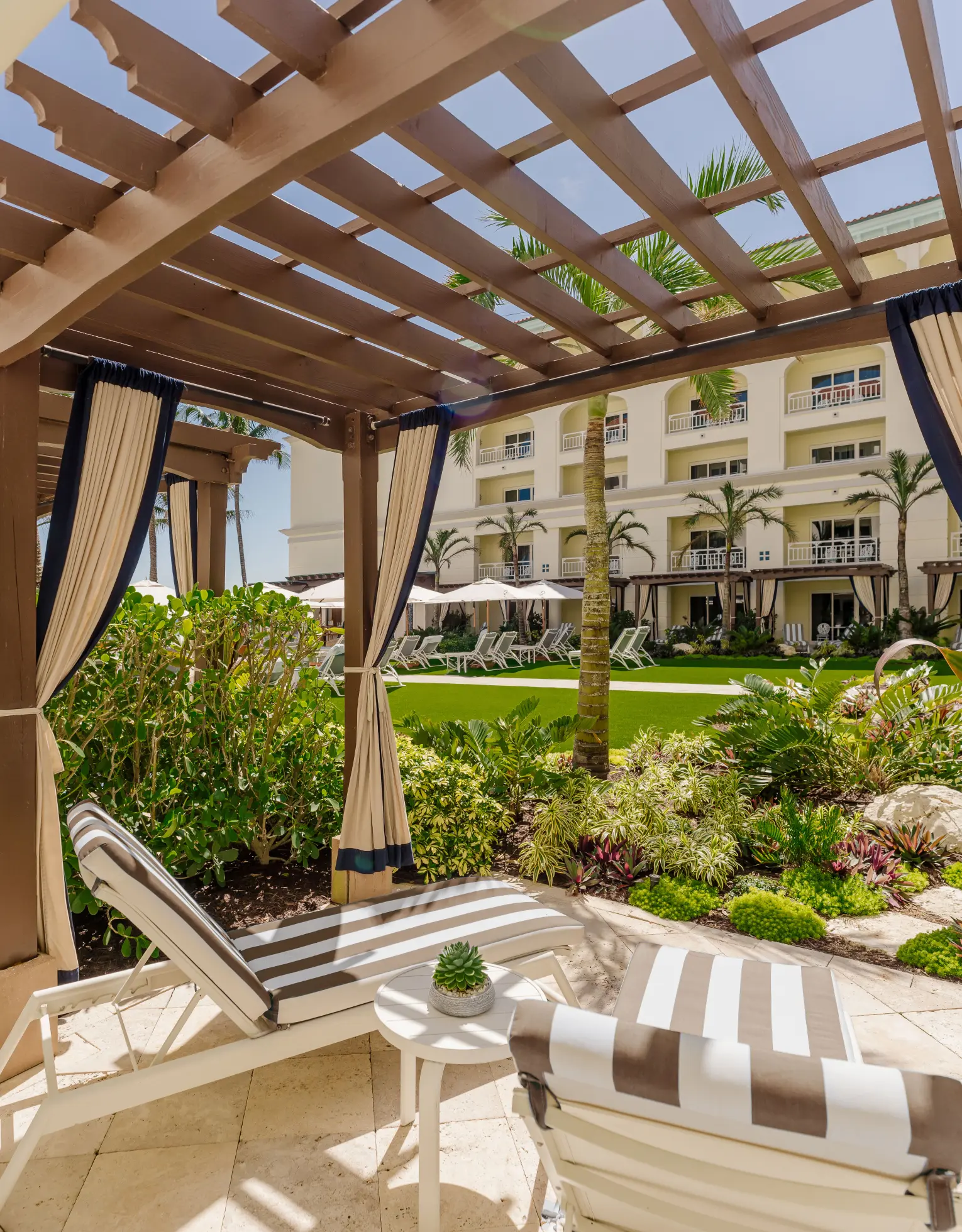 Pergola cabana with striped lounge chairs overlooking lush garden and resort courtyard with palm trees