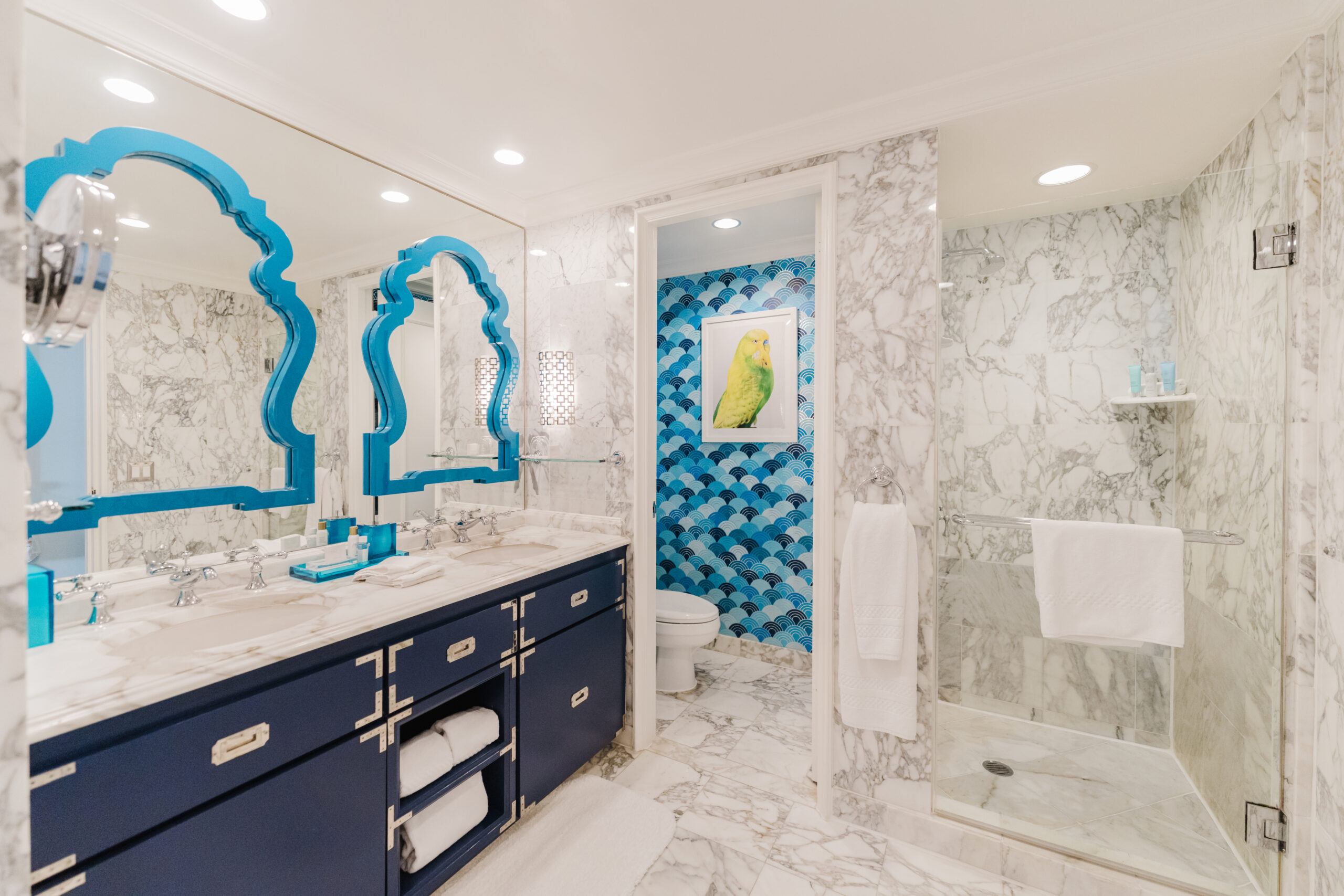 Marble bathroom with blue vanity, twin turquoise mirrors, glass shower, and patterned accent wall with parrot artwork