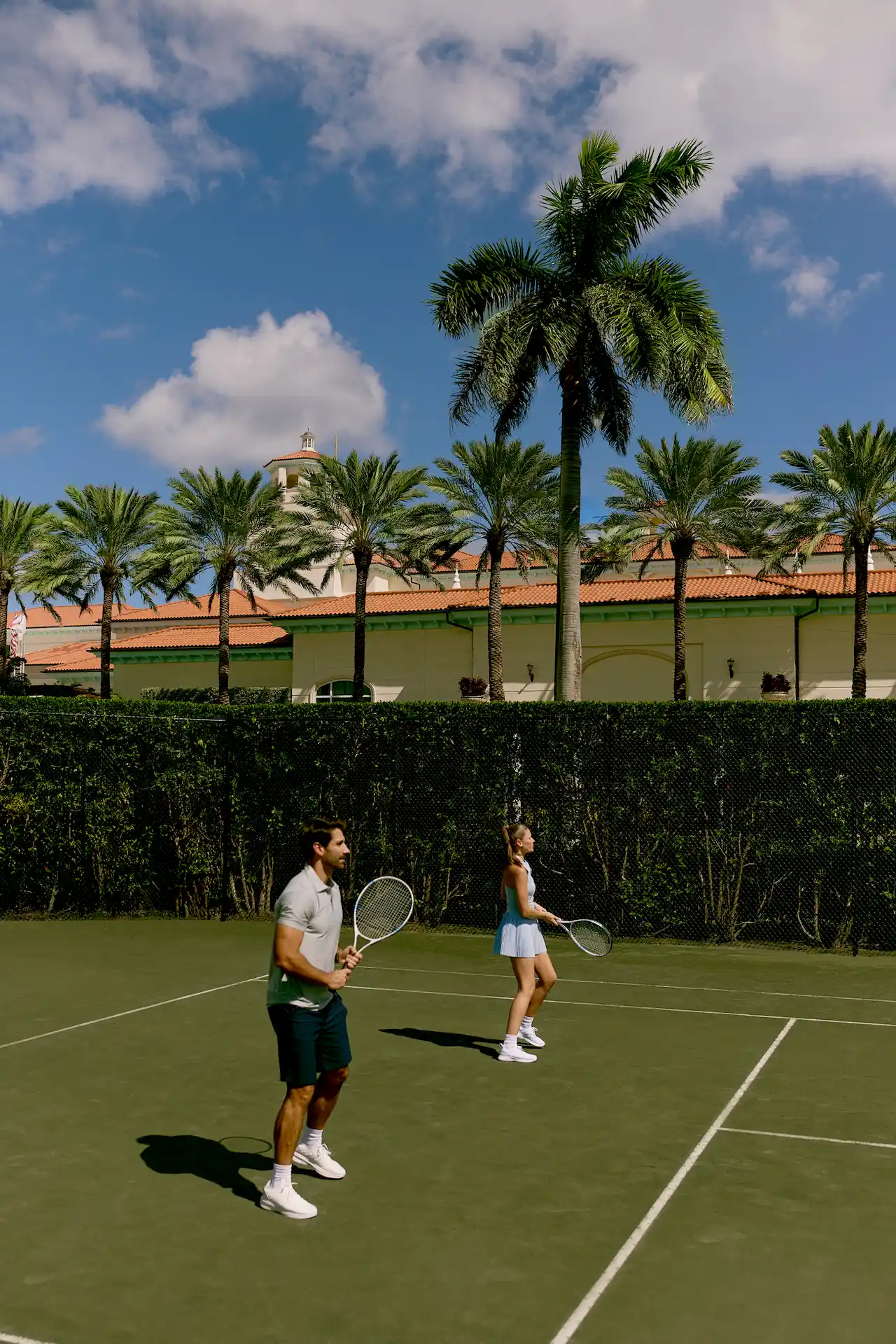 Couple walking by tennis court with palm trees and mediterranean style resort building behind