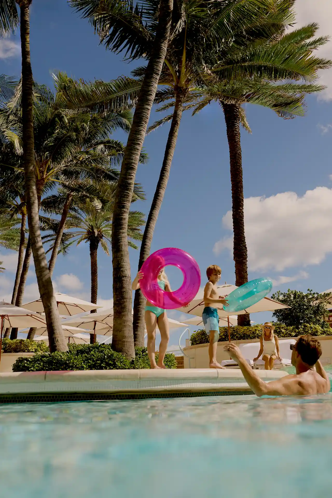 Family poolside moment with children holding colorful float rings as palm trees sway overhead in a sunny tropical setting