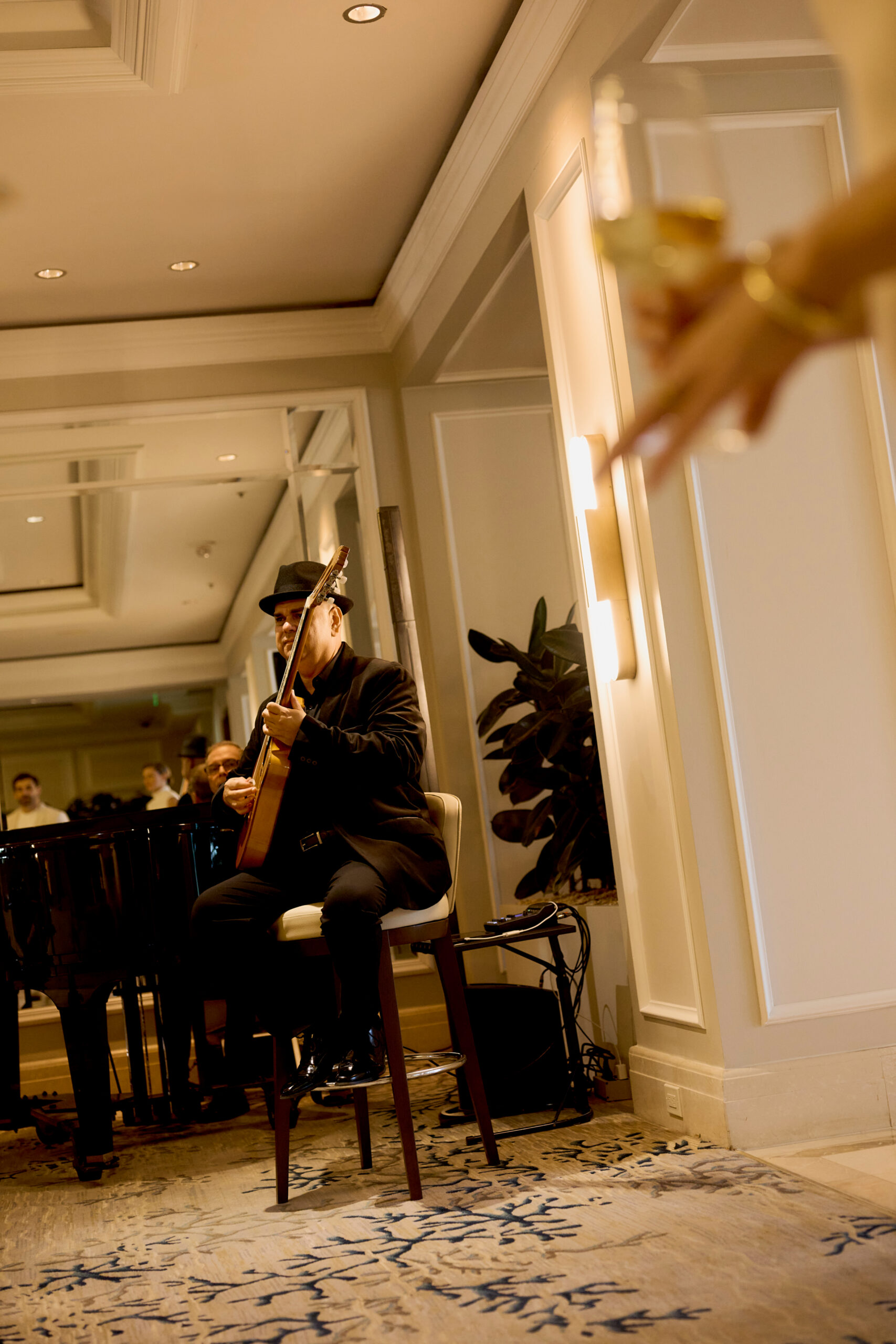 Musician in black suit and hat playing guitar beside a piano in an elegant indoor venue