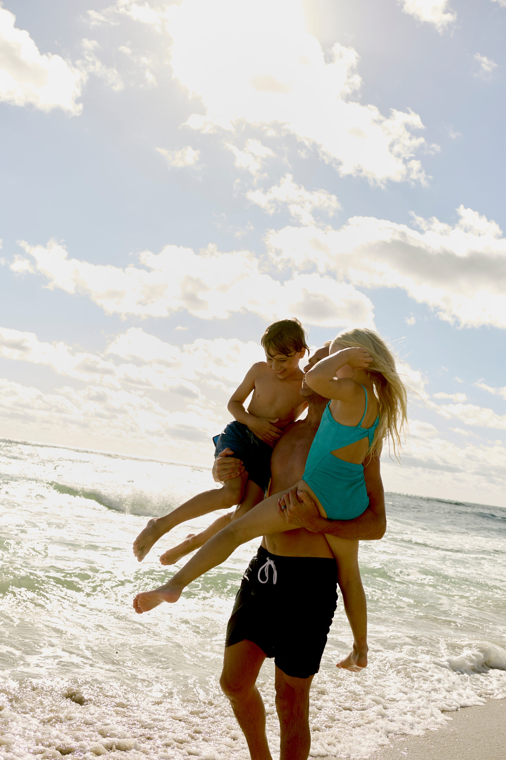 Father holding two children while standing in ocean waves under bright sunny sky