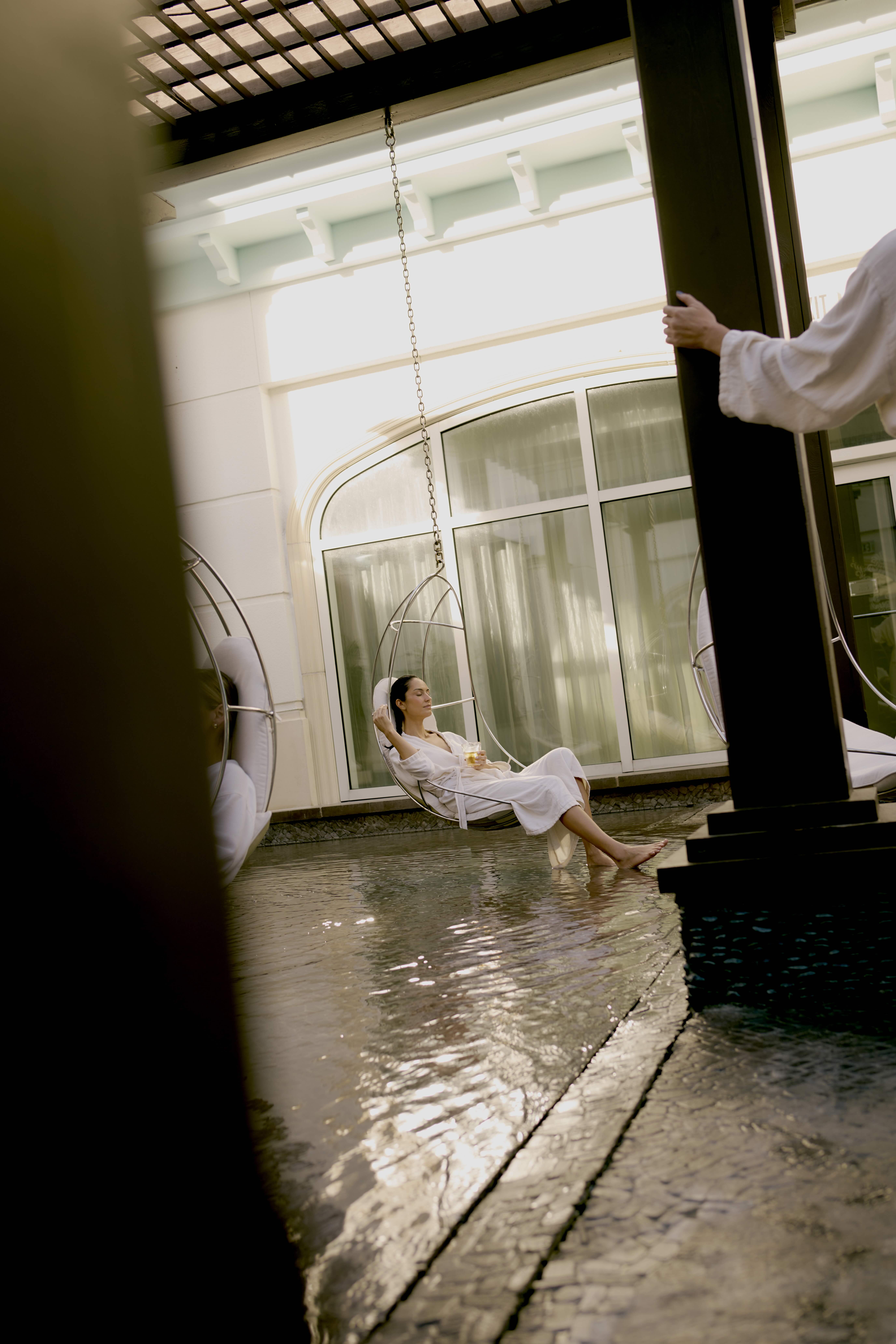 Woman in white robe relaxing on hanging chair above shallow water in luxury courtyard