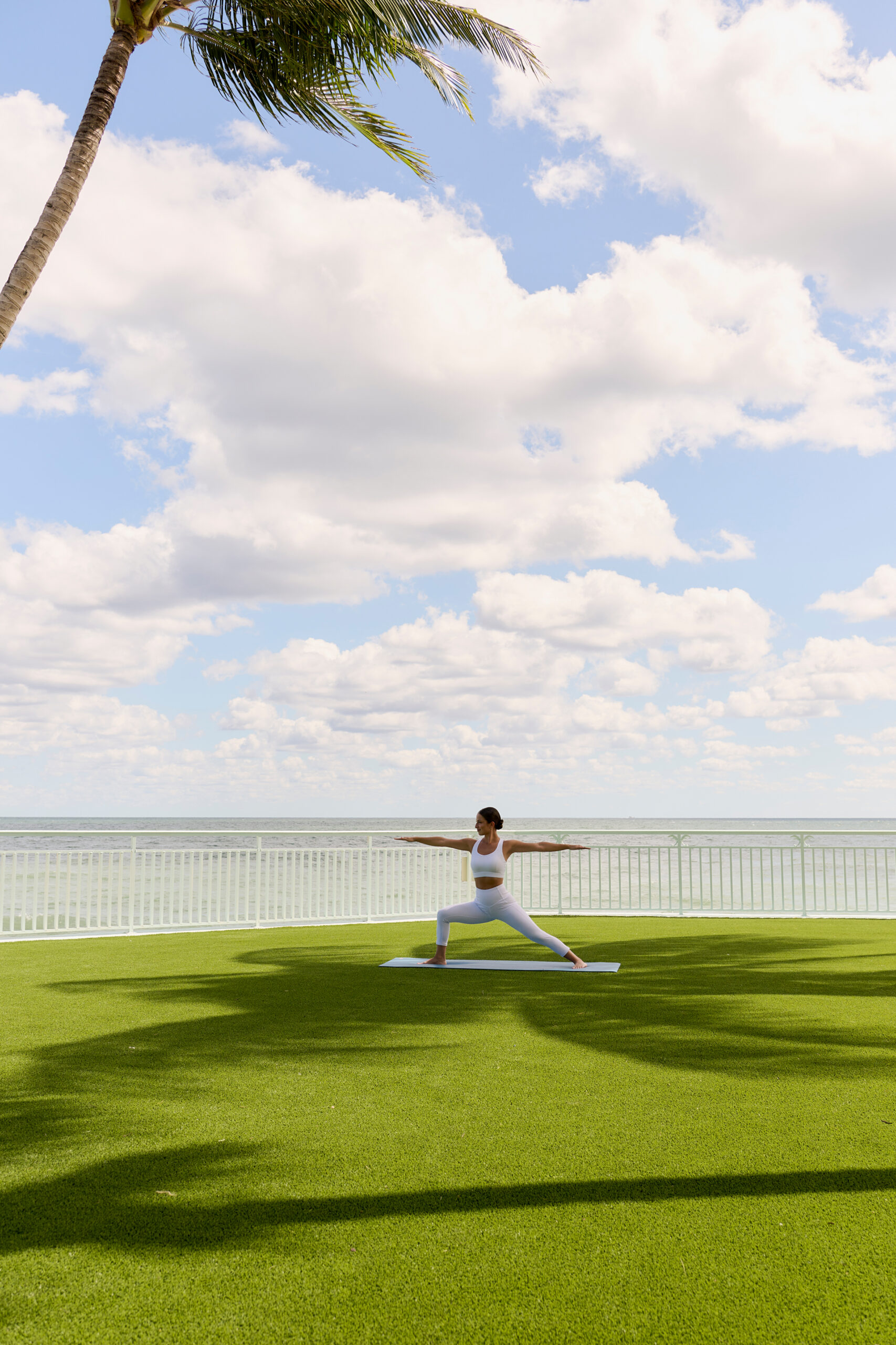 Woman practicing yoga on oceanfront lawn under palm tree with blue sky and white clouds