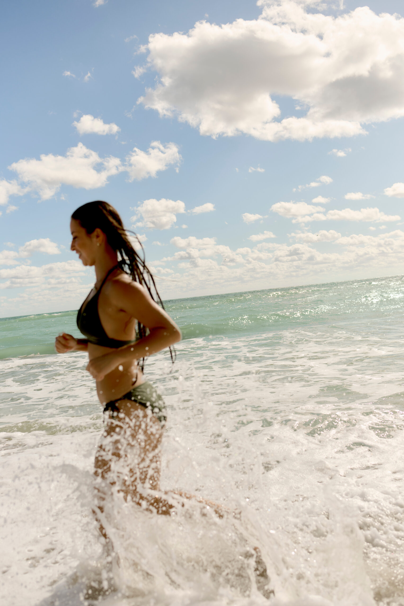 Woman in black bikini running through ocean waves under blue sky with scattered clouds