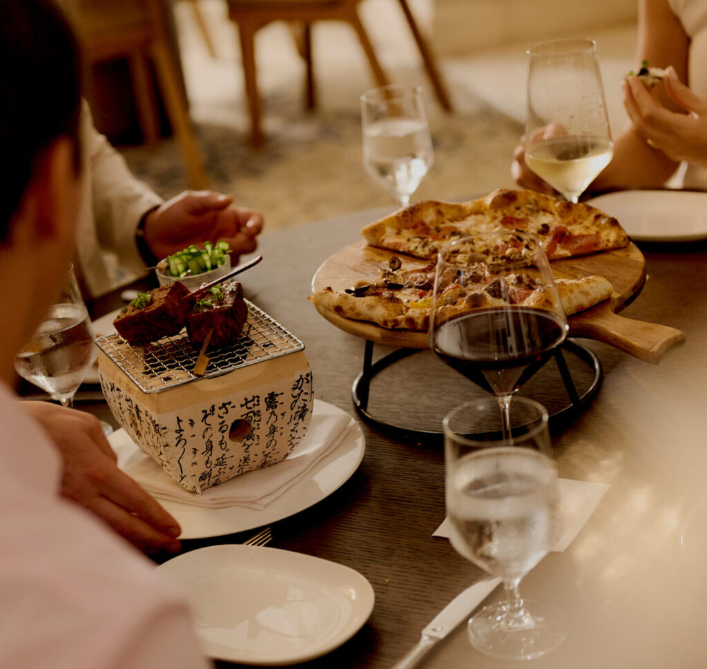 Friends sharing pizza and grilled skewers with wine at a warmly lit restaurant table