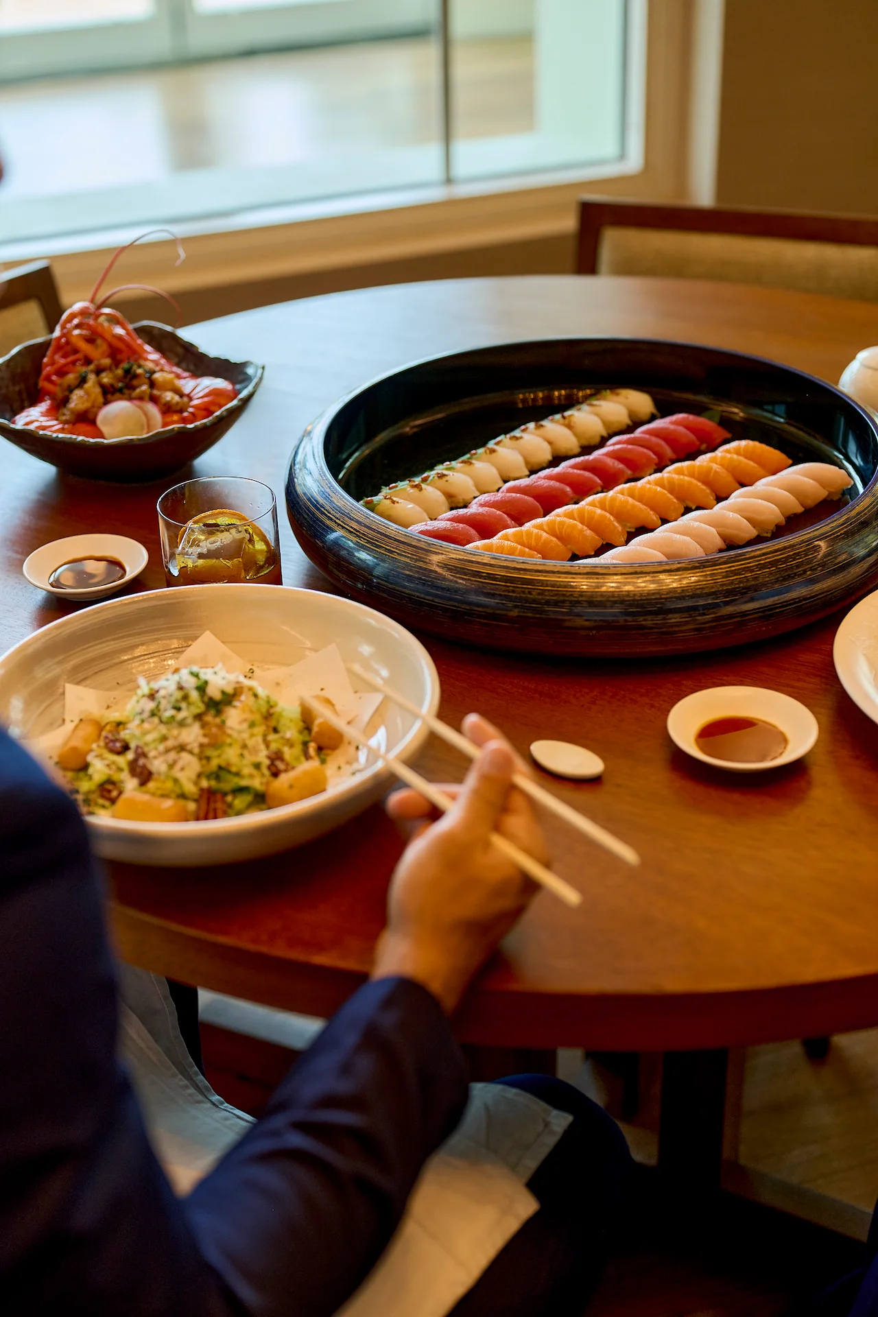 Person using chopsticks at table with assorted sushi platter, salad, and dipping sauces