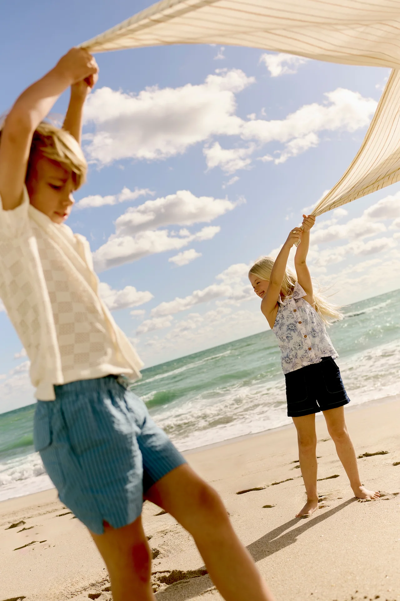 Two children holding fabric overhead and playing on sunny beach by the ocean