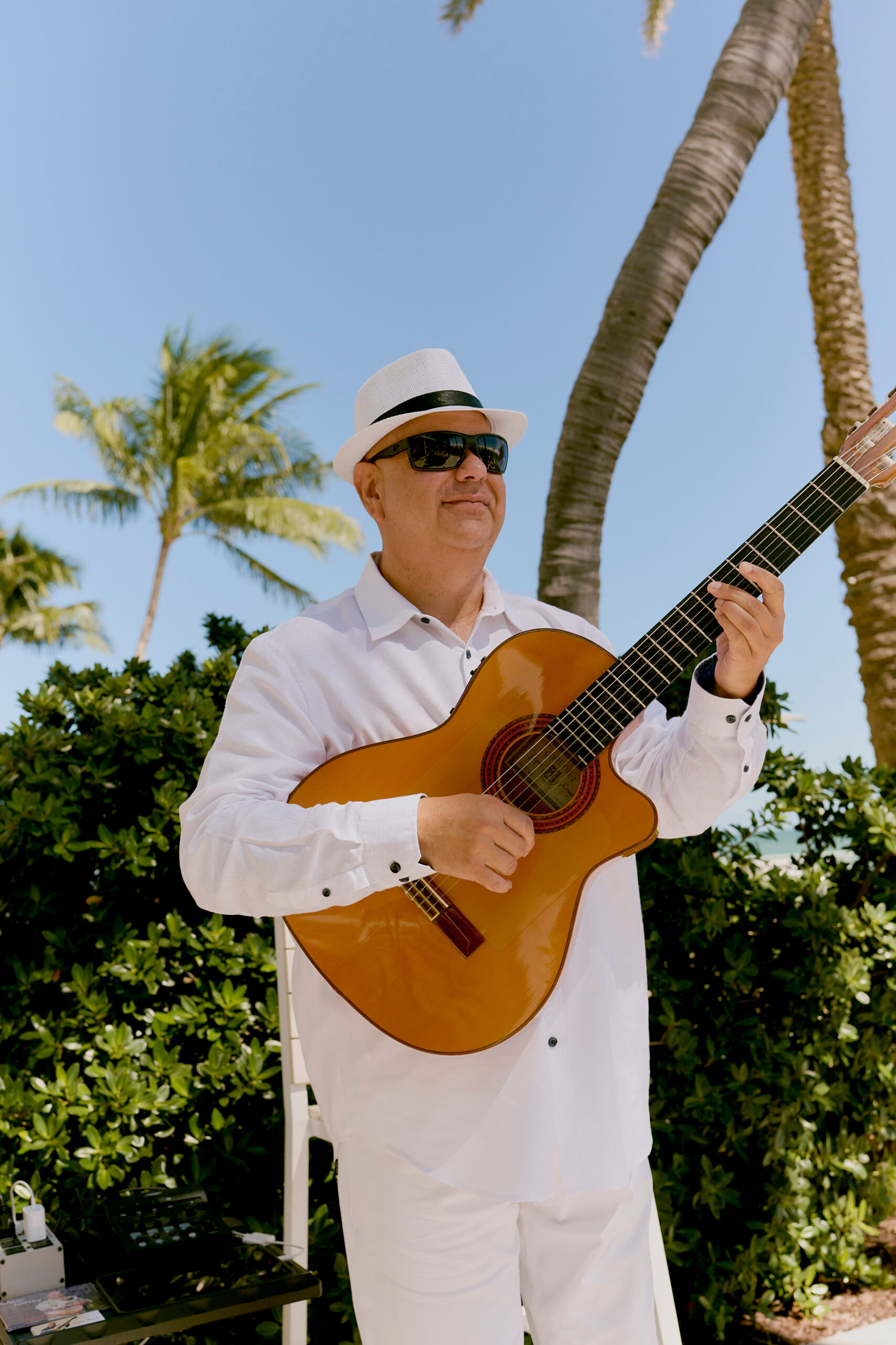 Man in white outfit and hat playing guitar outdoors with palm trees and blue sky