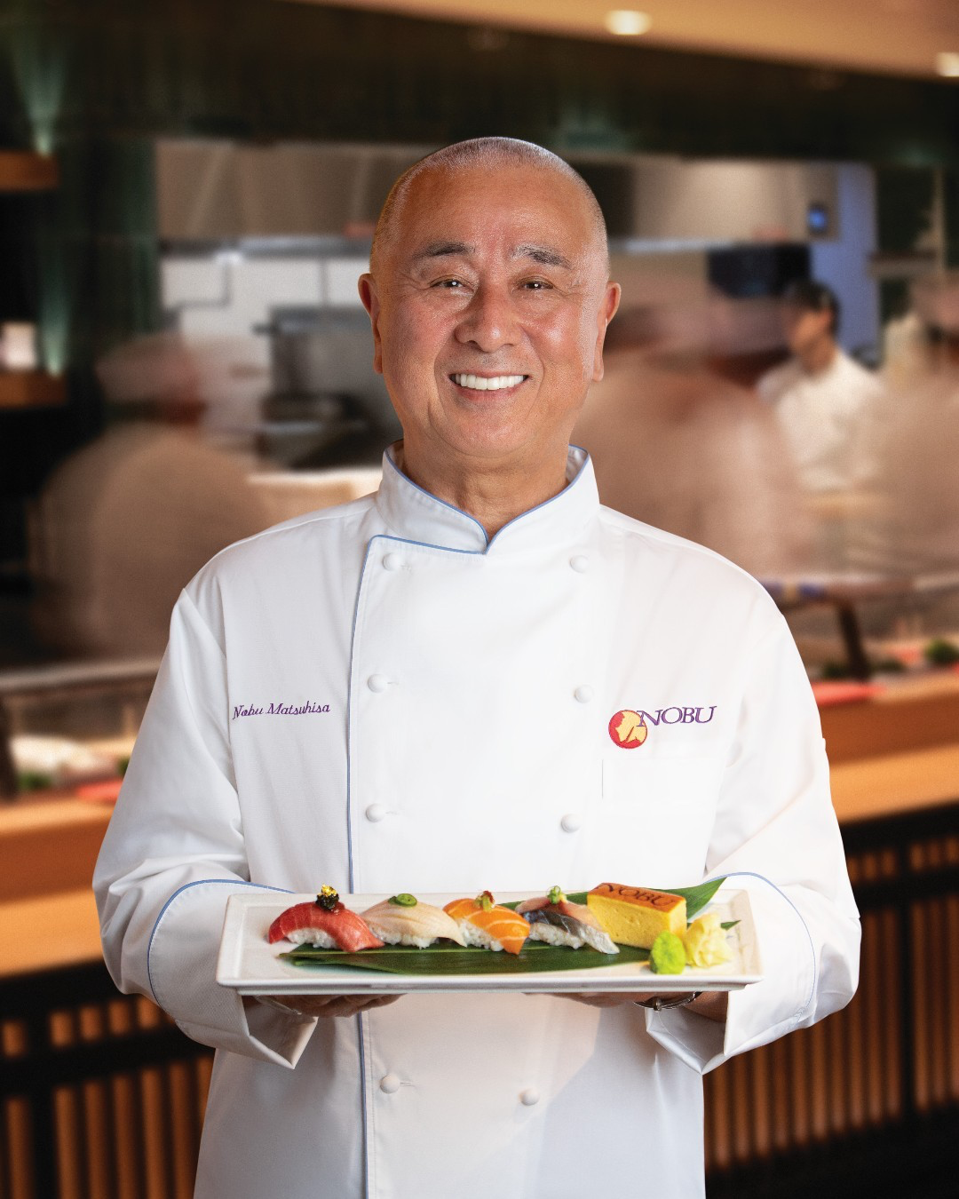 Smiling chef in white coat holding a platter of assorted sushi in a restaurant kitchen setting