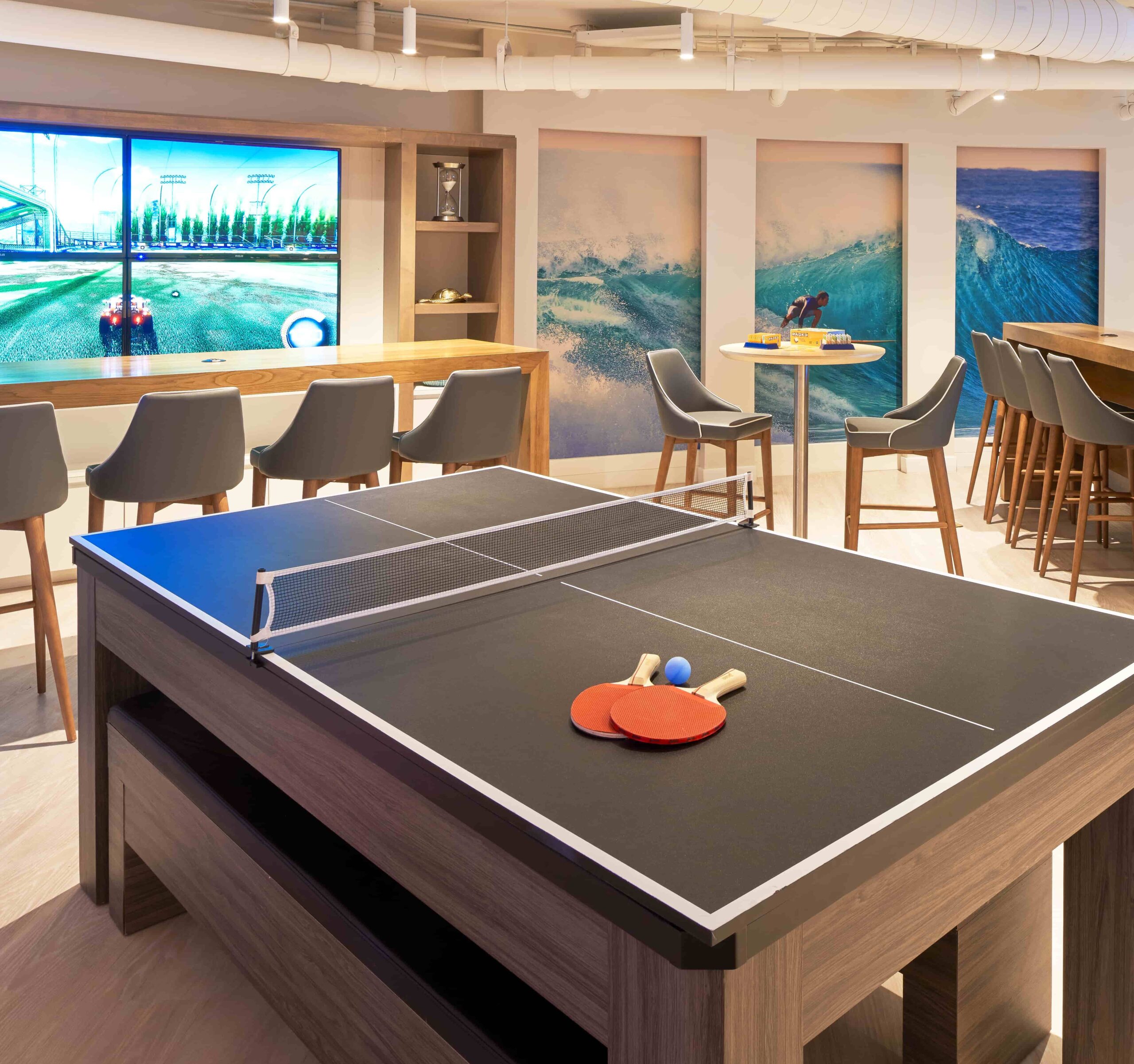 Modern office lounge with ping pong table, bar seating, large gaming screen, and ocean wave wall art