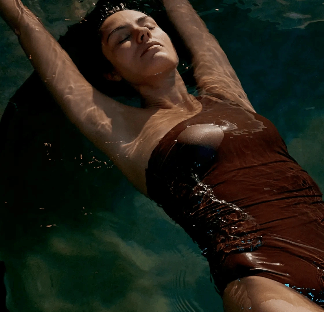 Woman in brown swimsuit floating peacefully in sunlit pool with arms extended and eyes closed