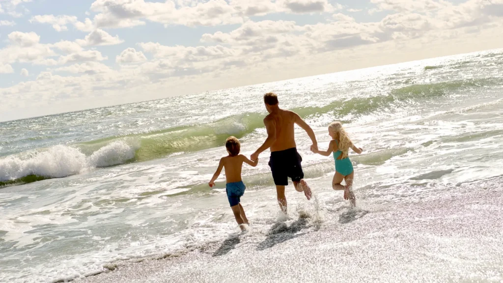Father holding hands with two children running into ocean waves on sunny beach
