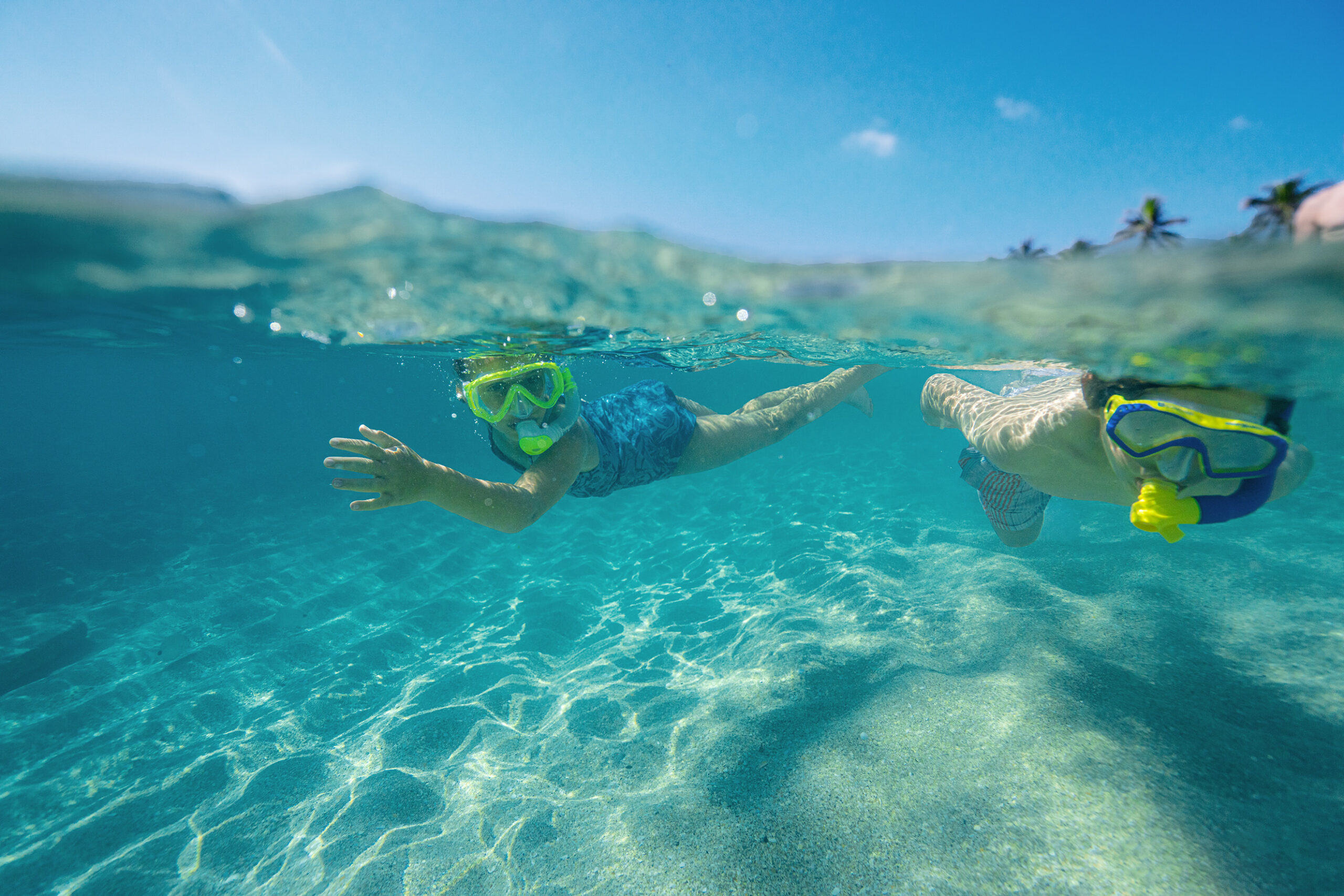 Two children snorkeling in clear turquoise ocean water above sandy seabed near a tropical shore