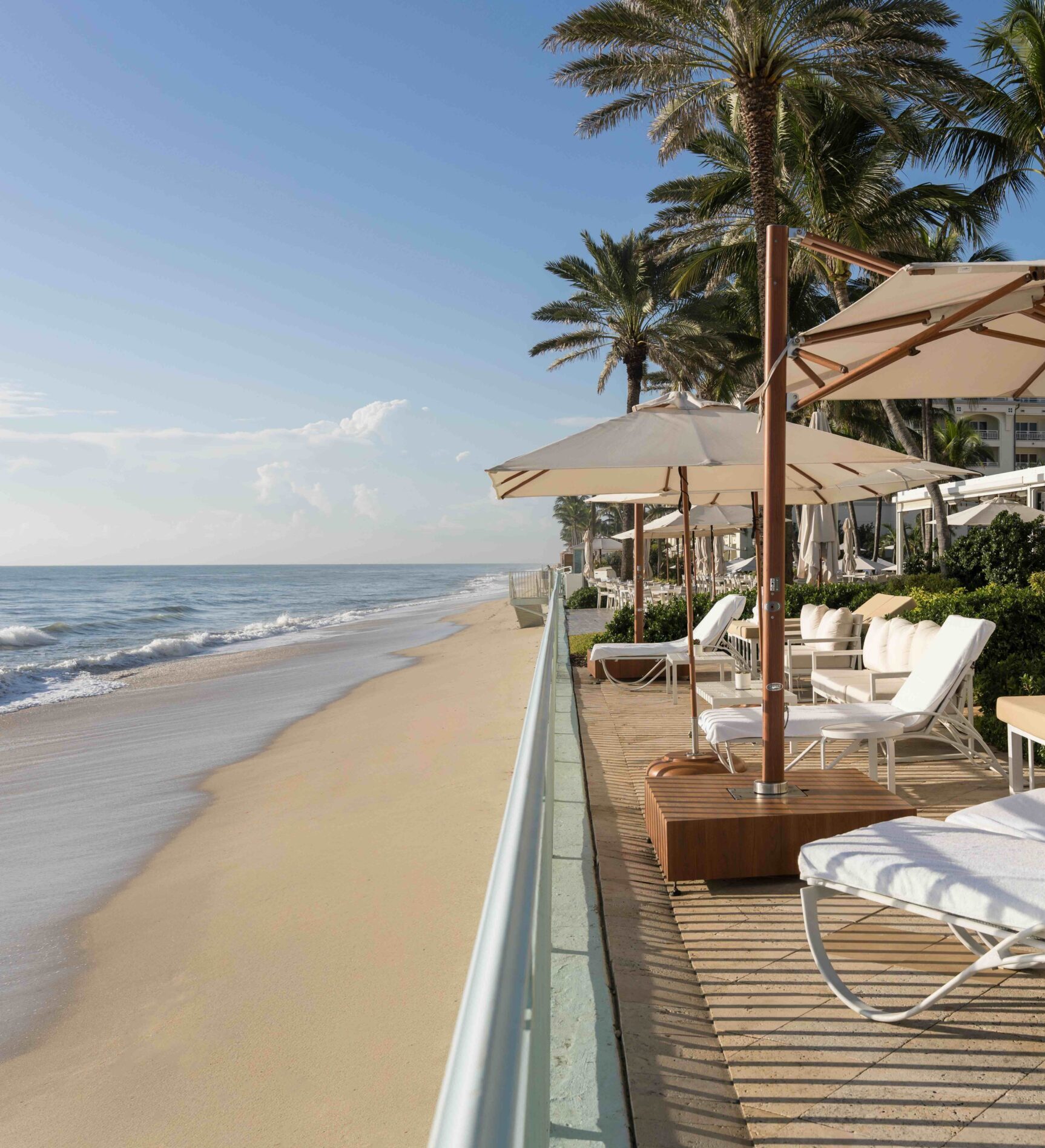 Beachfront lounge area with white chairs, umbrellas, and palm trees.