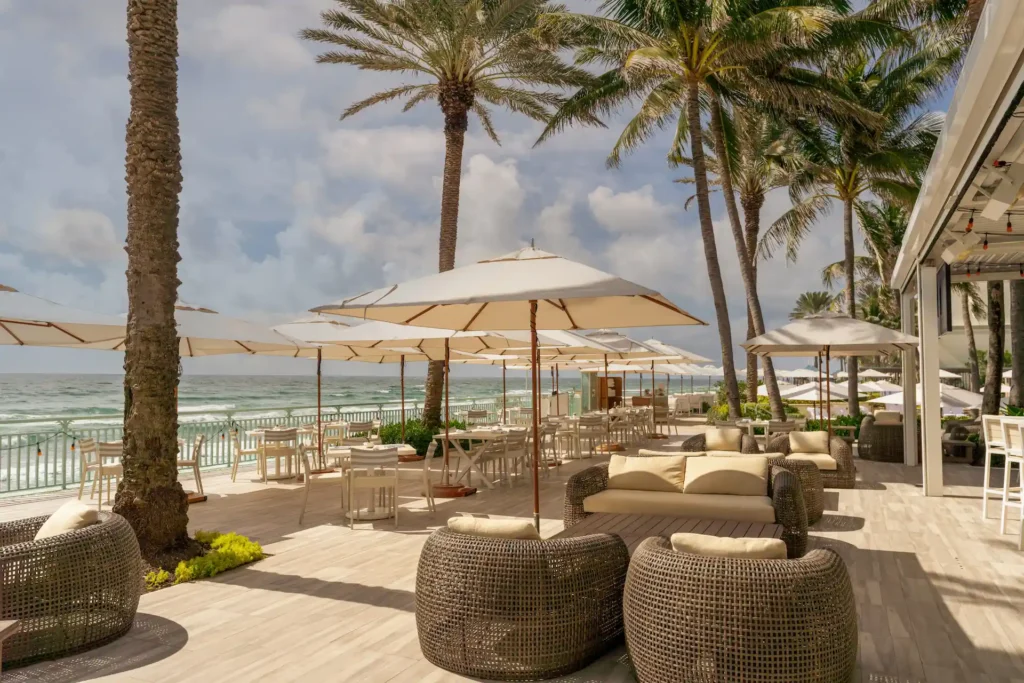 Oceanfront lounge at breeze ocean kitchen with white umbrellas, palm trees and relaxed coastal seating at eau palm beach