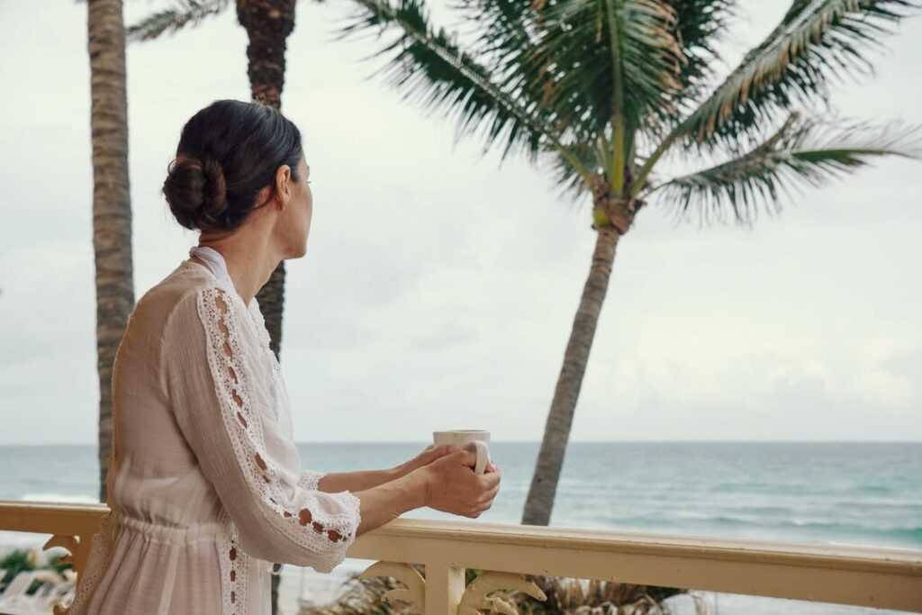 Woman holding coffee on seaside balcony with palm trees and ocean view