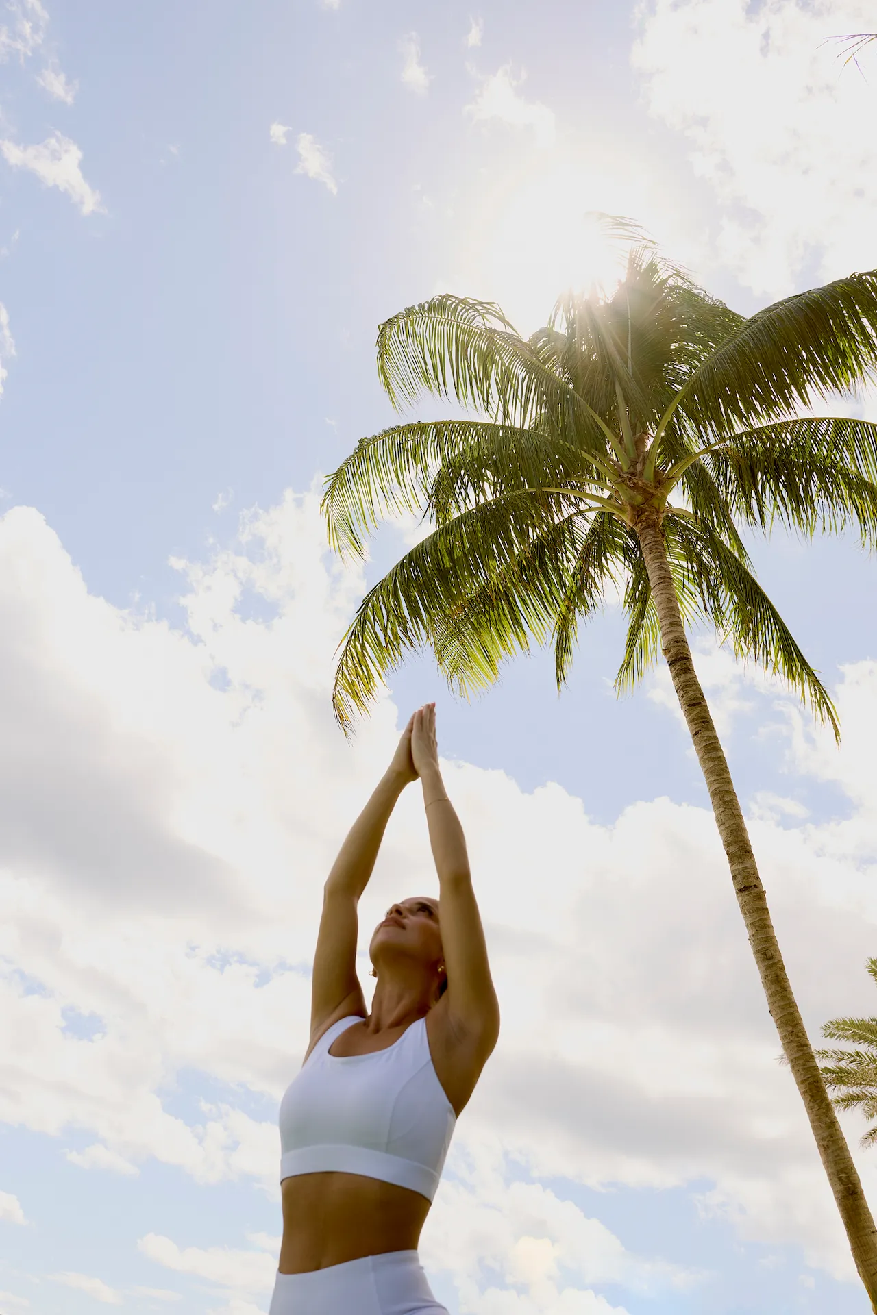Woman practicing yoga under palm tree with sunlit sky and scattered clouds