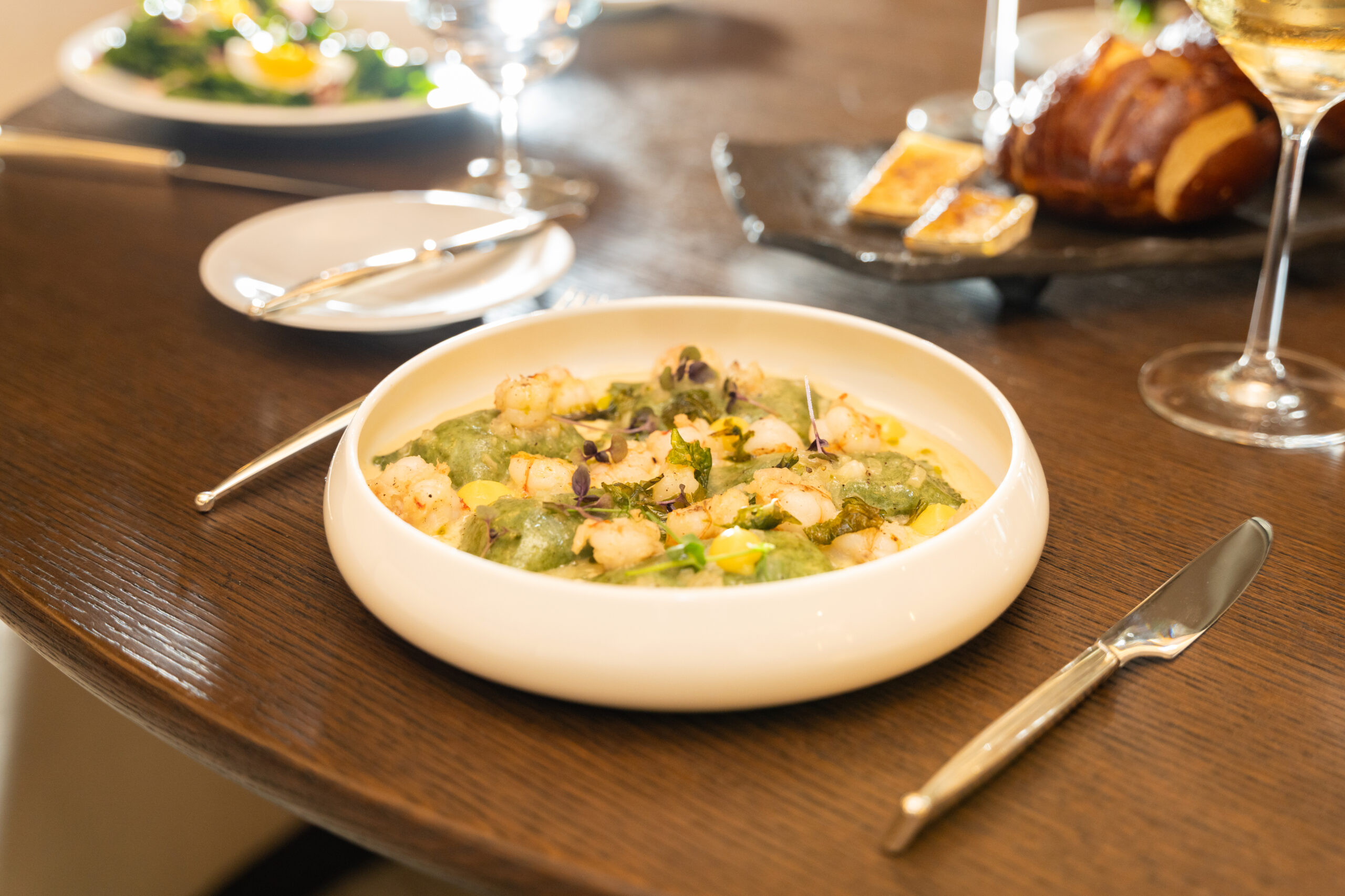 Shrimp and green ravioli in creamy sauce on a wooden table with wine glasses and bread in the background