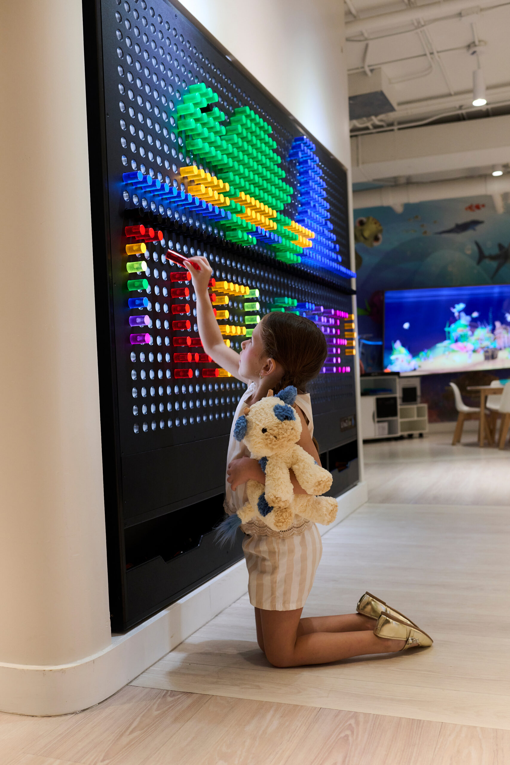 Young girl holding a teddy bear while placing colorful pegs on a large interactive wall display