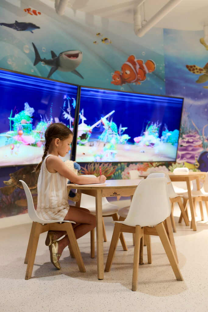 Young girl drawing at small table in colorful ocean-themed playroom with animated sea life mural