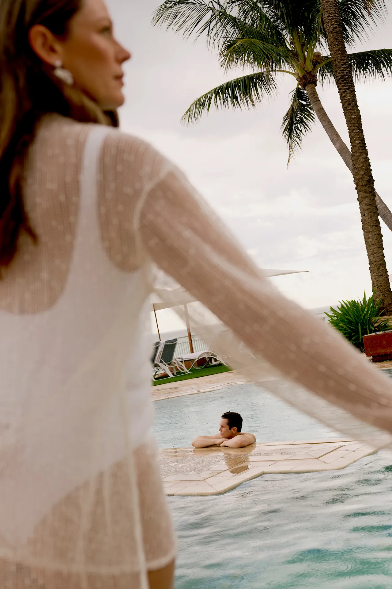 Person in a light cover‑up by the pool while another relaxes in the water under swaying palm trees