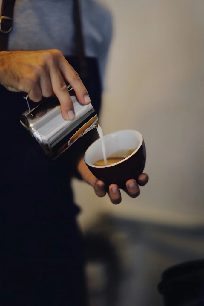 Barista pouring steamed milk from metal pitcher into cup of coffee