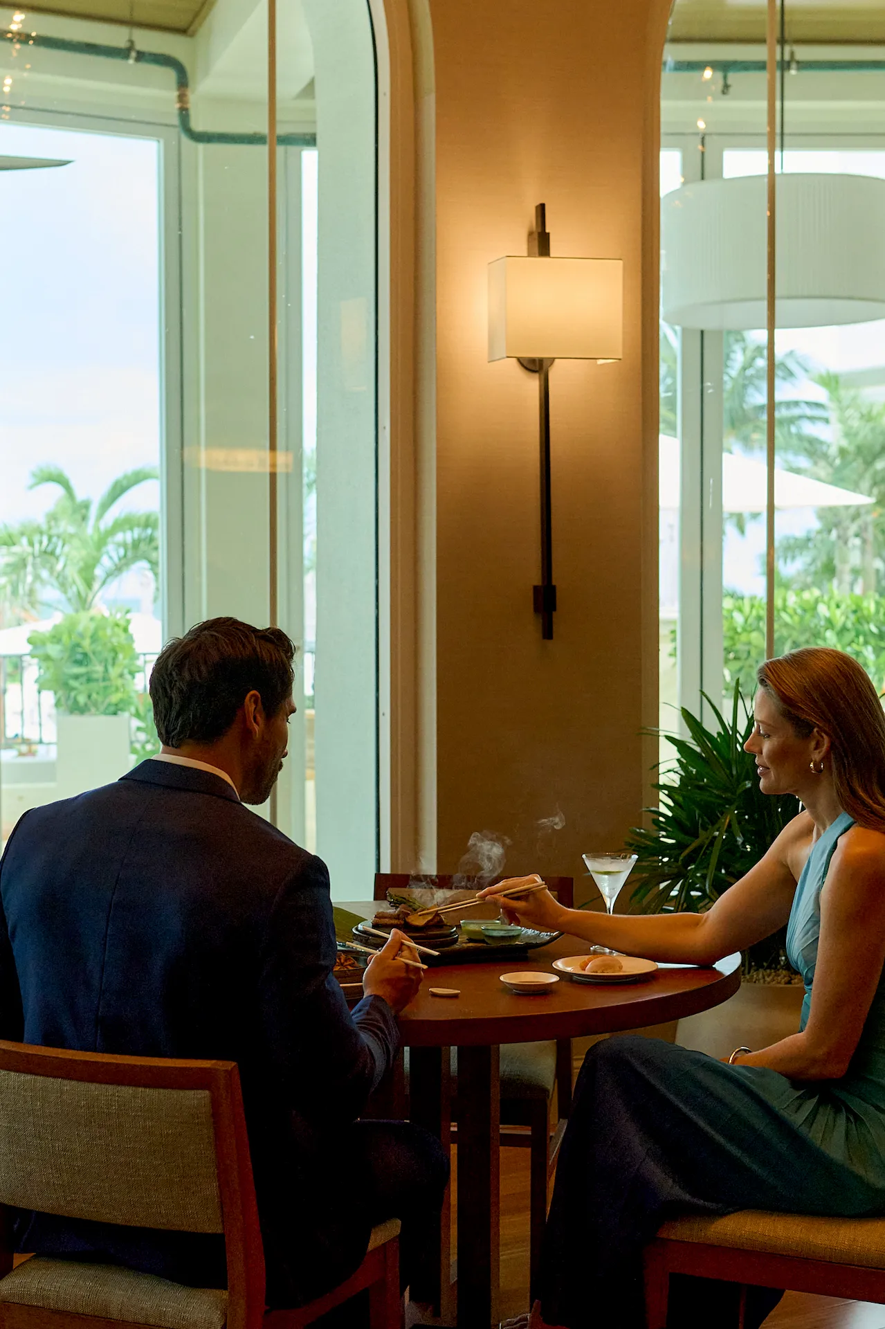 Fine dining moment with two guests enjoying a refined meal by large windows overlooking tropical greenery