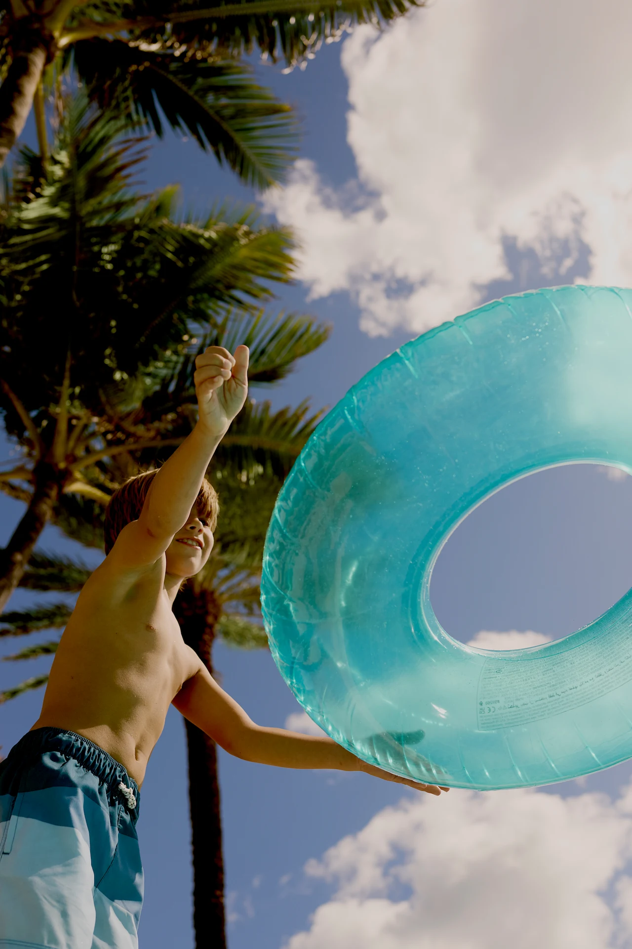 Smiling boy holding blue inflatable swim ring under palm trees and sunny sky