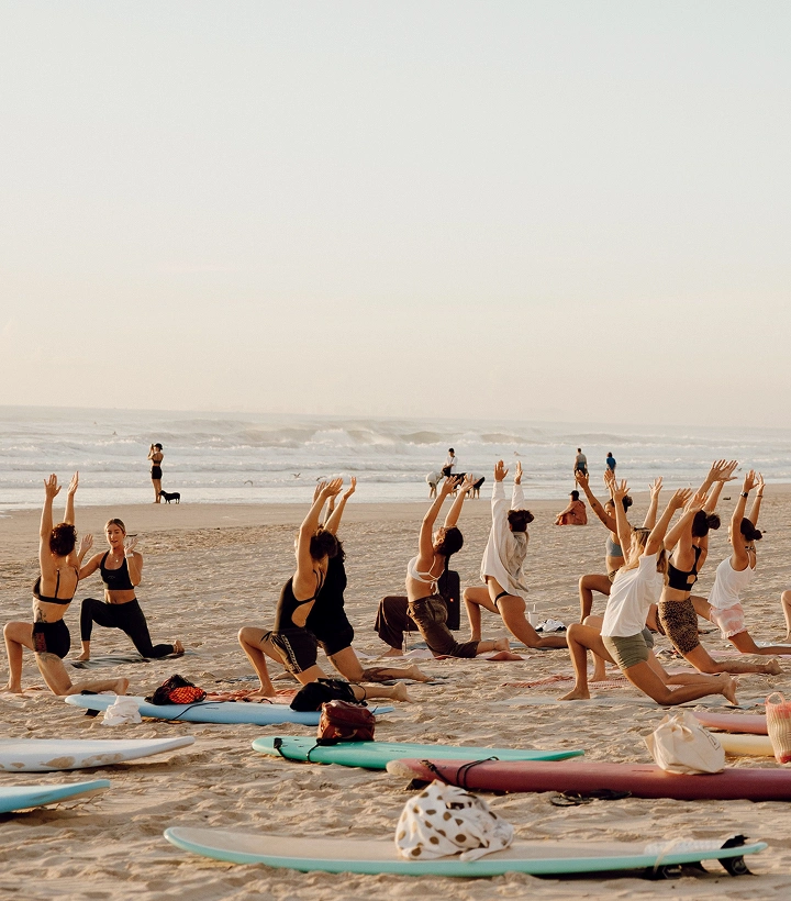 Group practicing yoga lunges on sandy beach at sunrise, with surfboards and ocean waves in the background