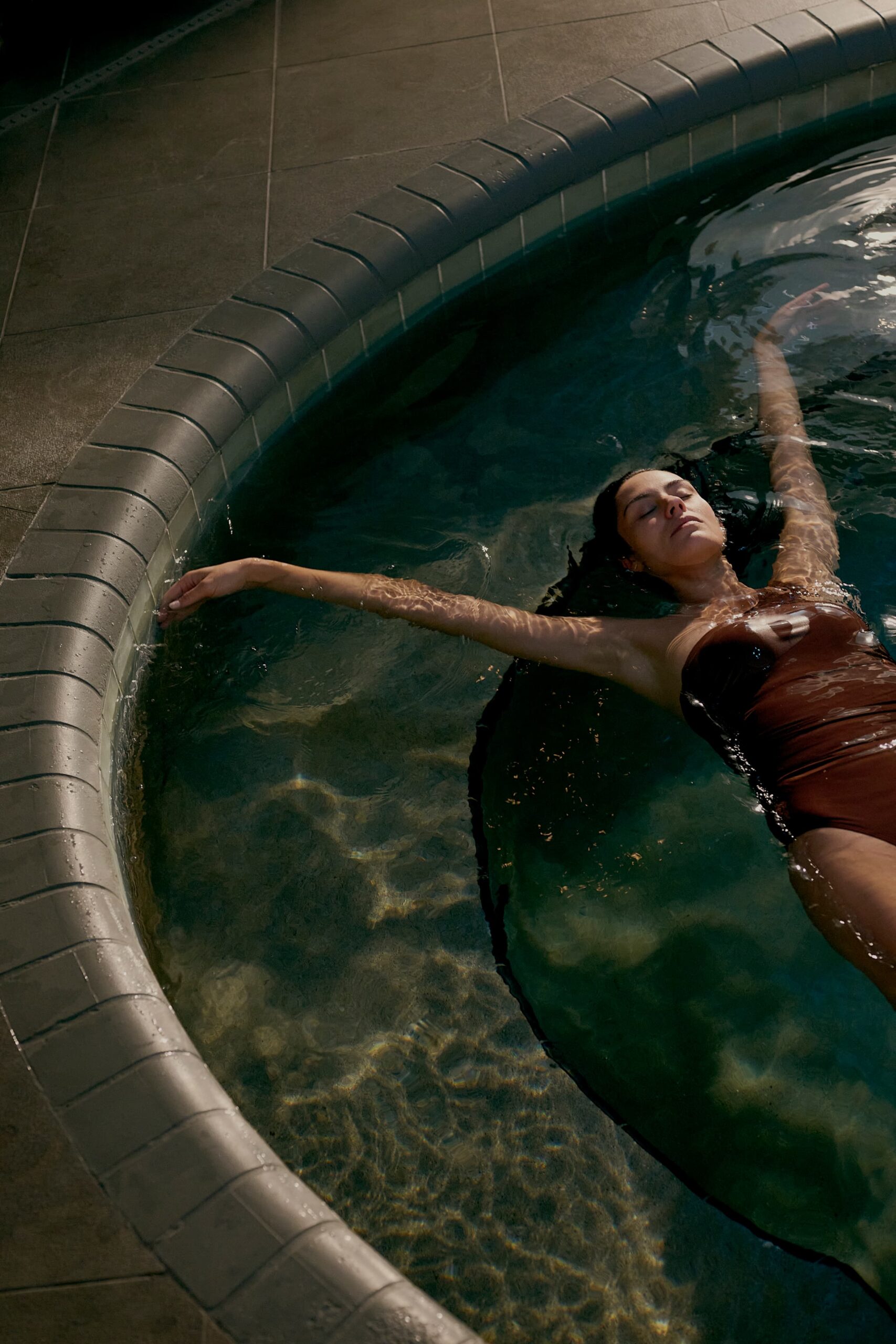 Woman floating on her back in a curved pool with eyes closed, relaxing in sunlight