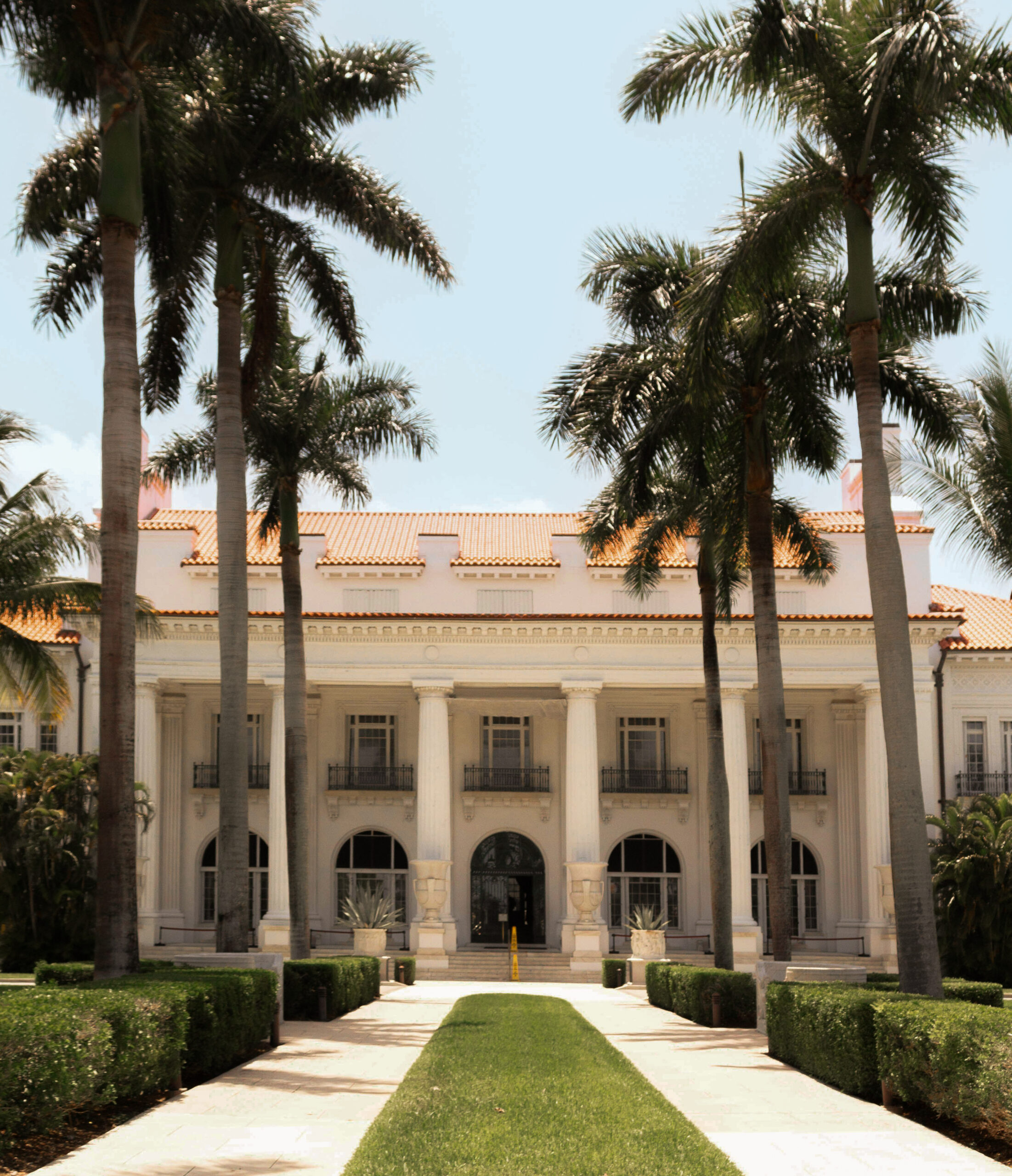 Grand white mansion with tall columns and palm trees lining walkway under clear blue sky