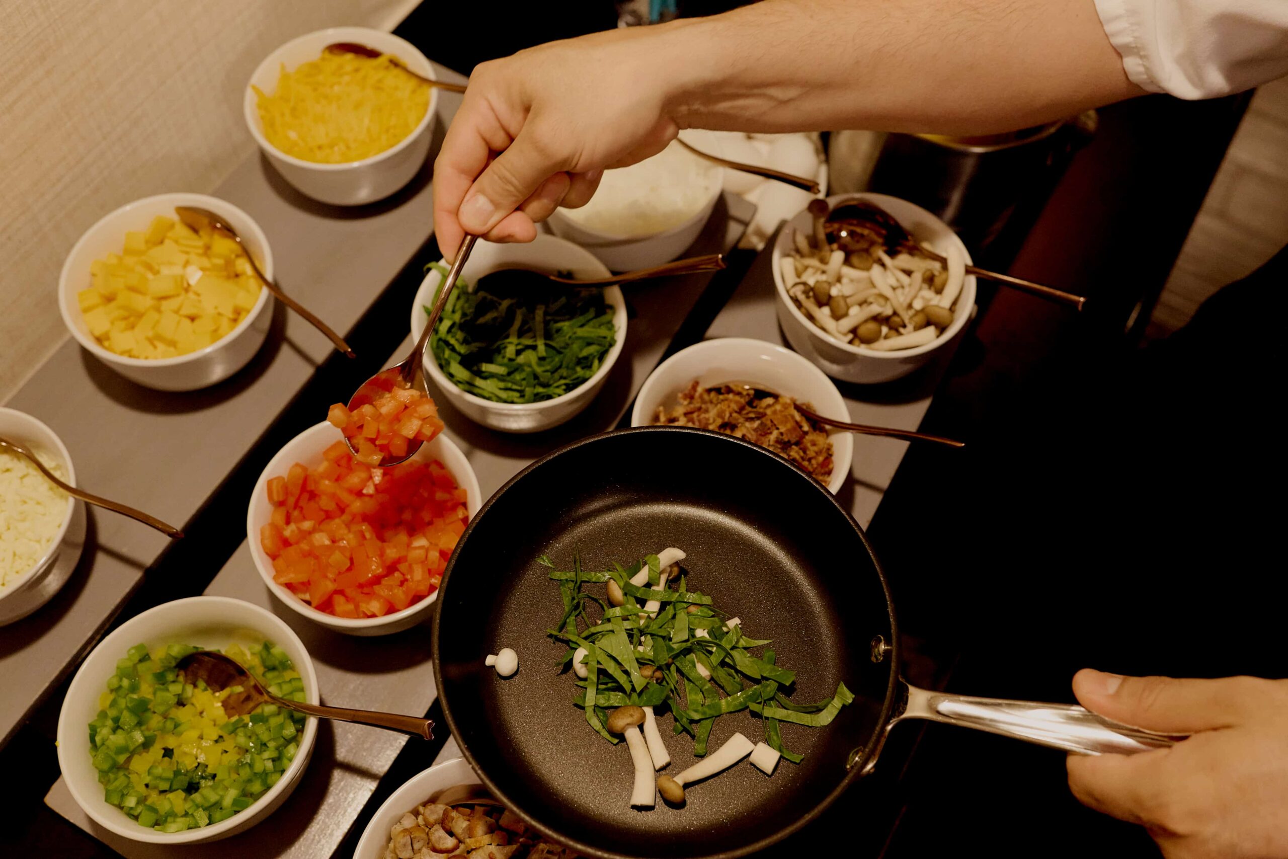 Hand adding diced tomatoes to pan with greens and mushrooms, surrounded by bowls of chopped ingredients