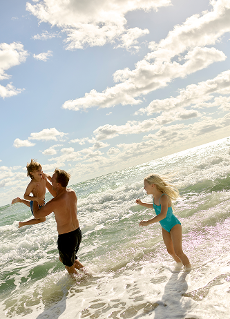 Father lifting young child while another child runs through ocean waves under a sunny sky