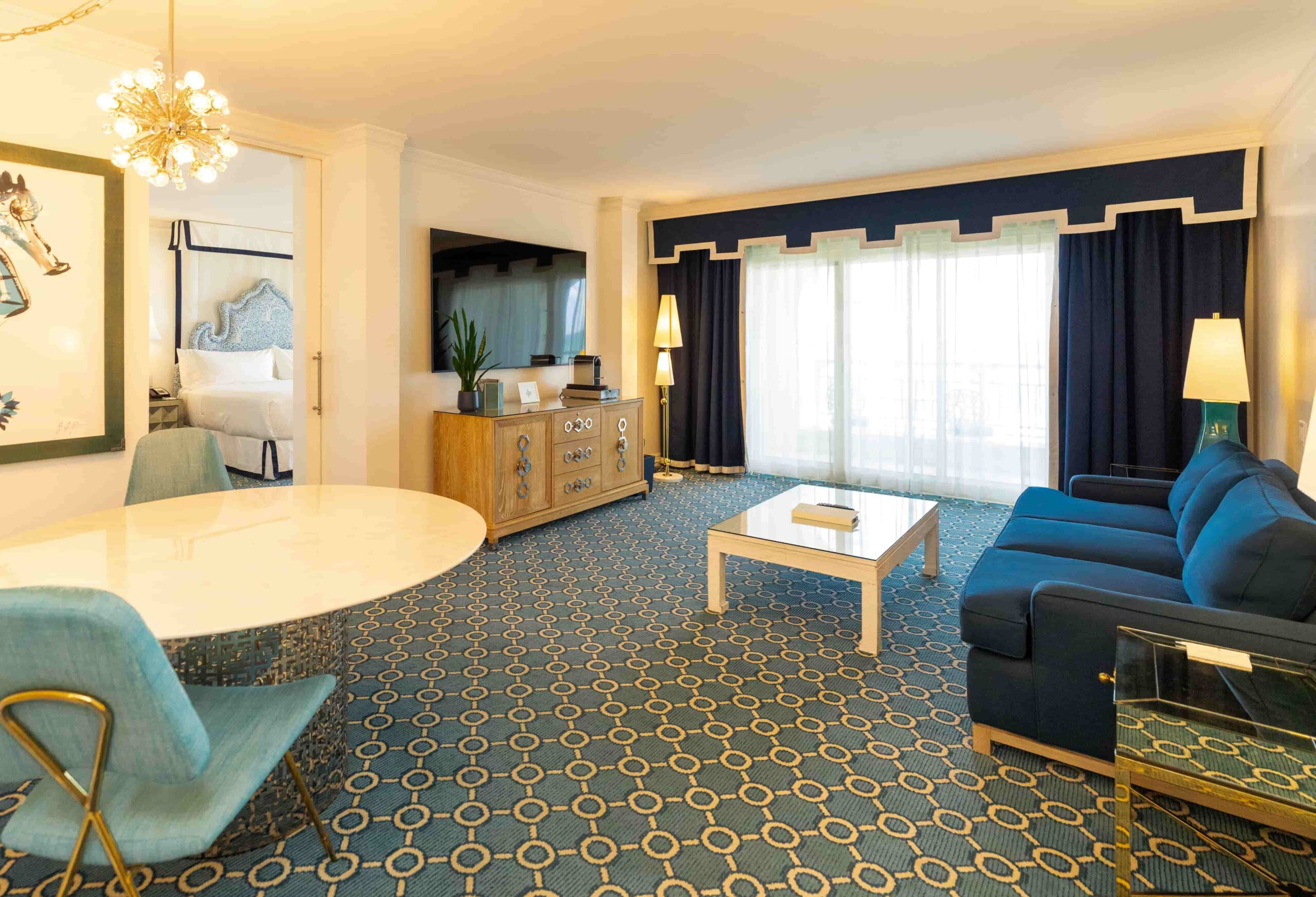 Spacious hotel suite with blue sofa, patterned carpet, large windows, and separate bedroom area