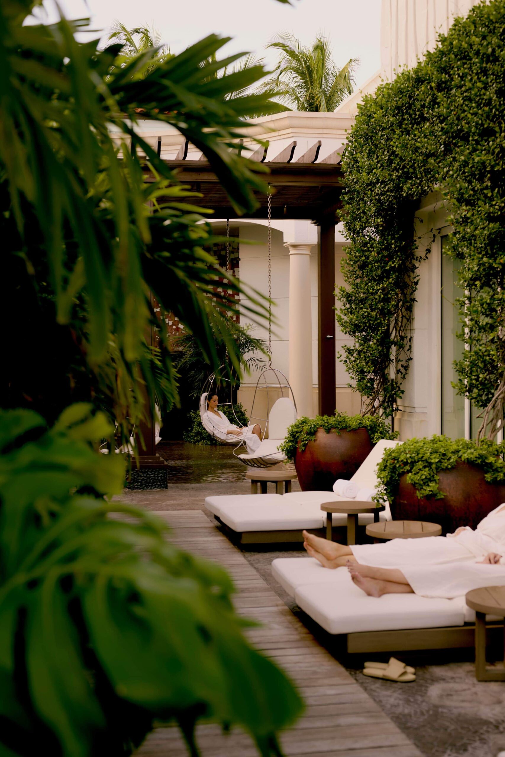Lush tropical spa courtyard with lounge chairs and woman relaxing in hanging chair under pergola