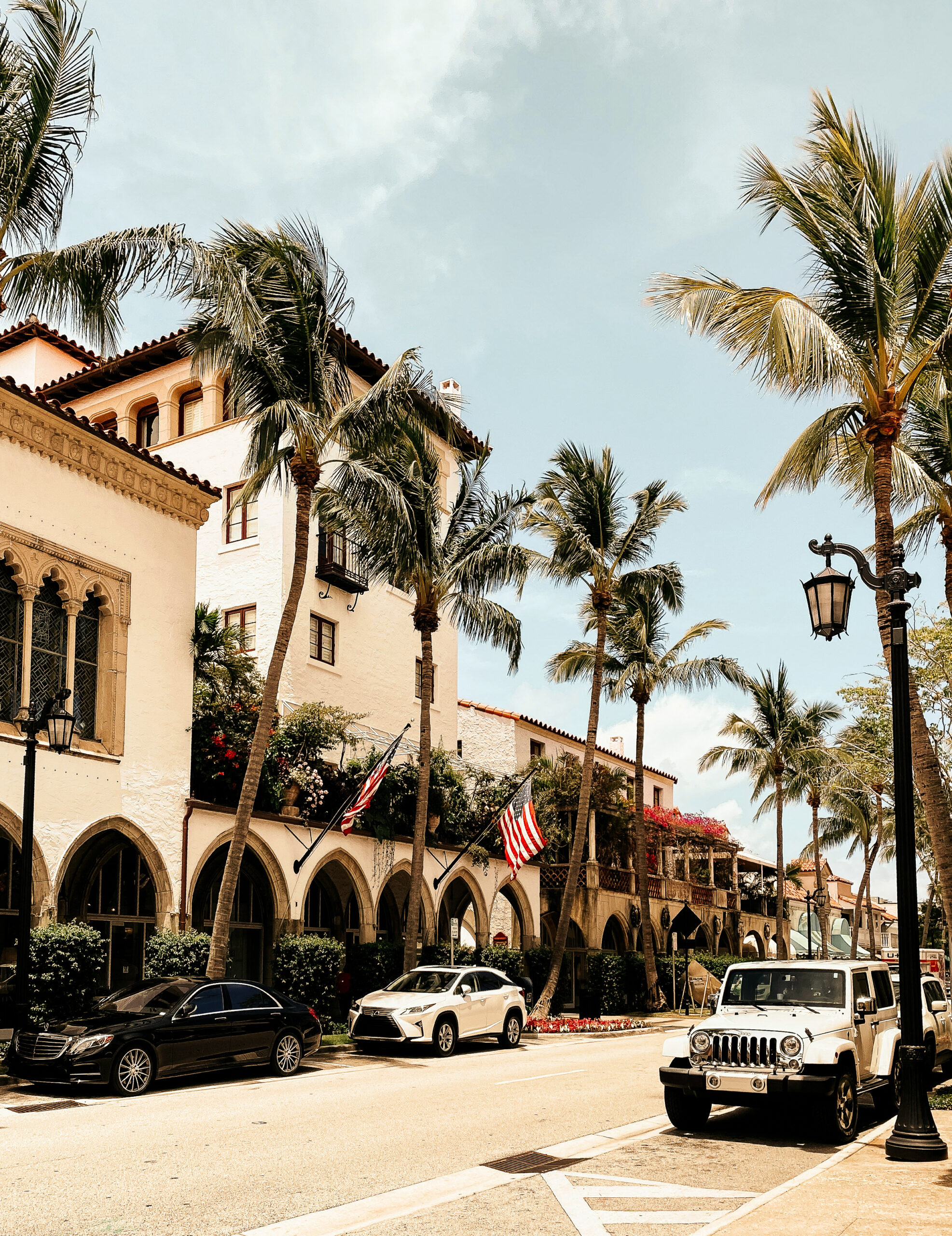Palm lined street with mediterranean style buildings, parked cars, and american flags under blue sky
