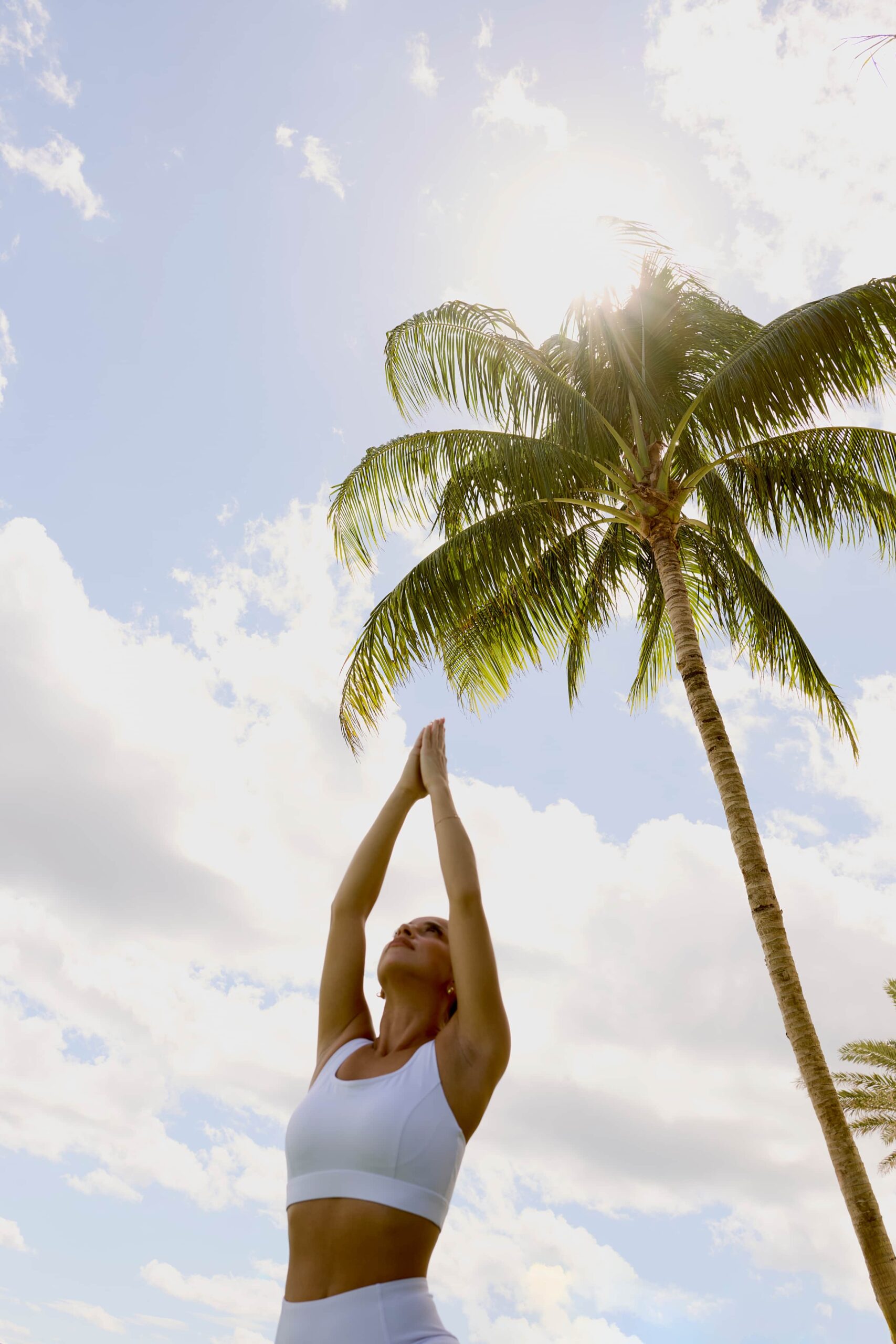Woman in white activewear practicing yoga with arms raised under a palm tree in bright sunlight