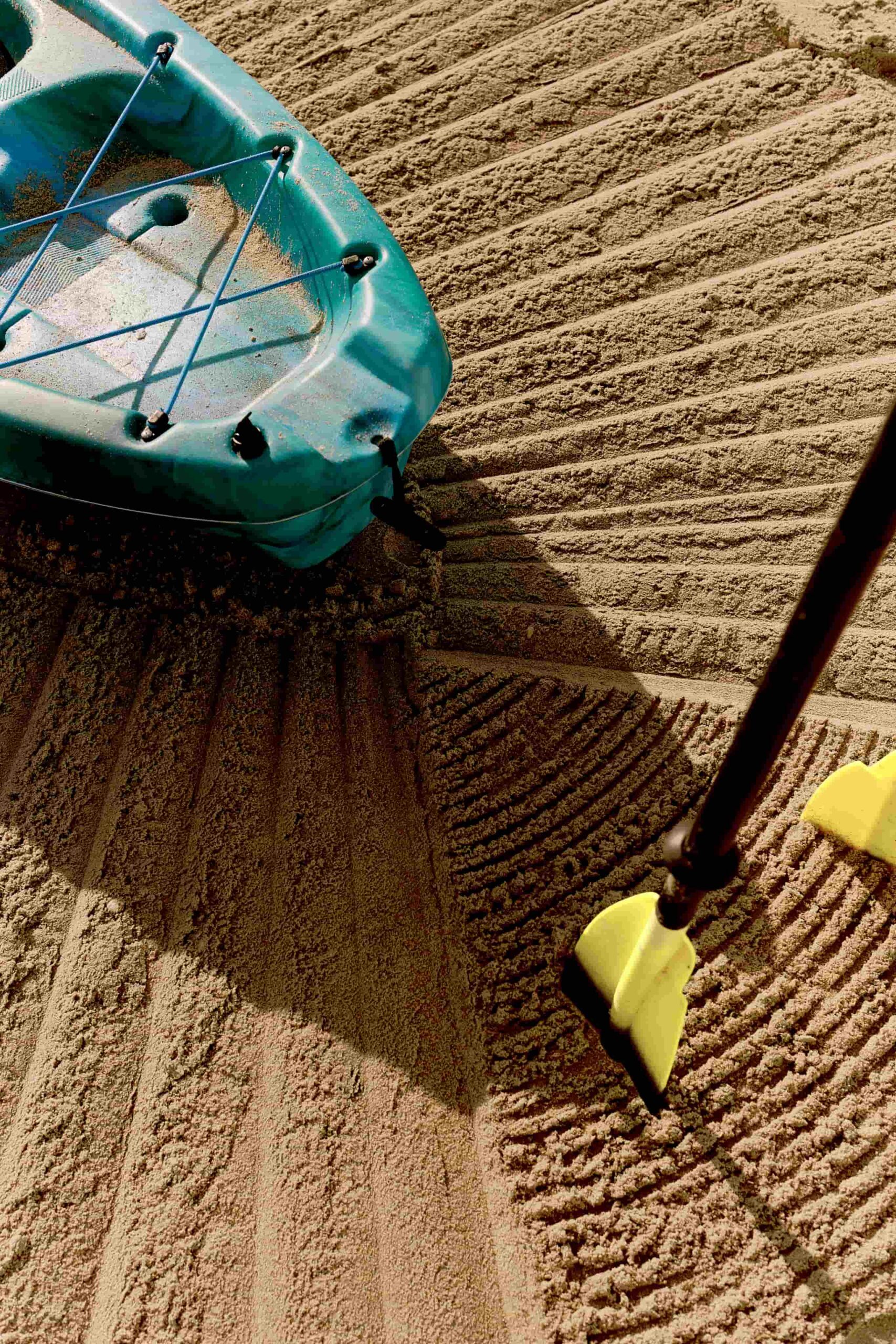 Turquoise kayak and paddle resting on raked sand beach with textured patterns in sunlight