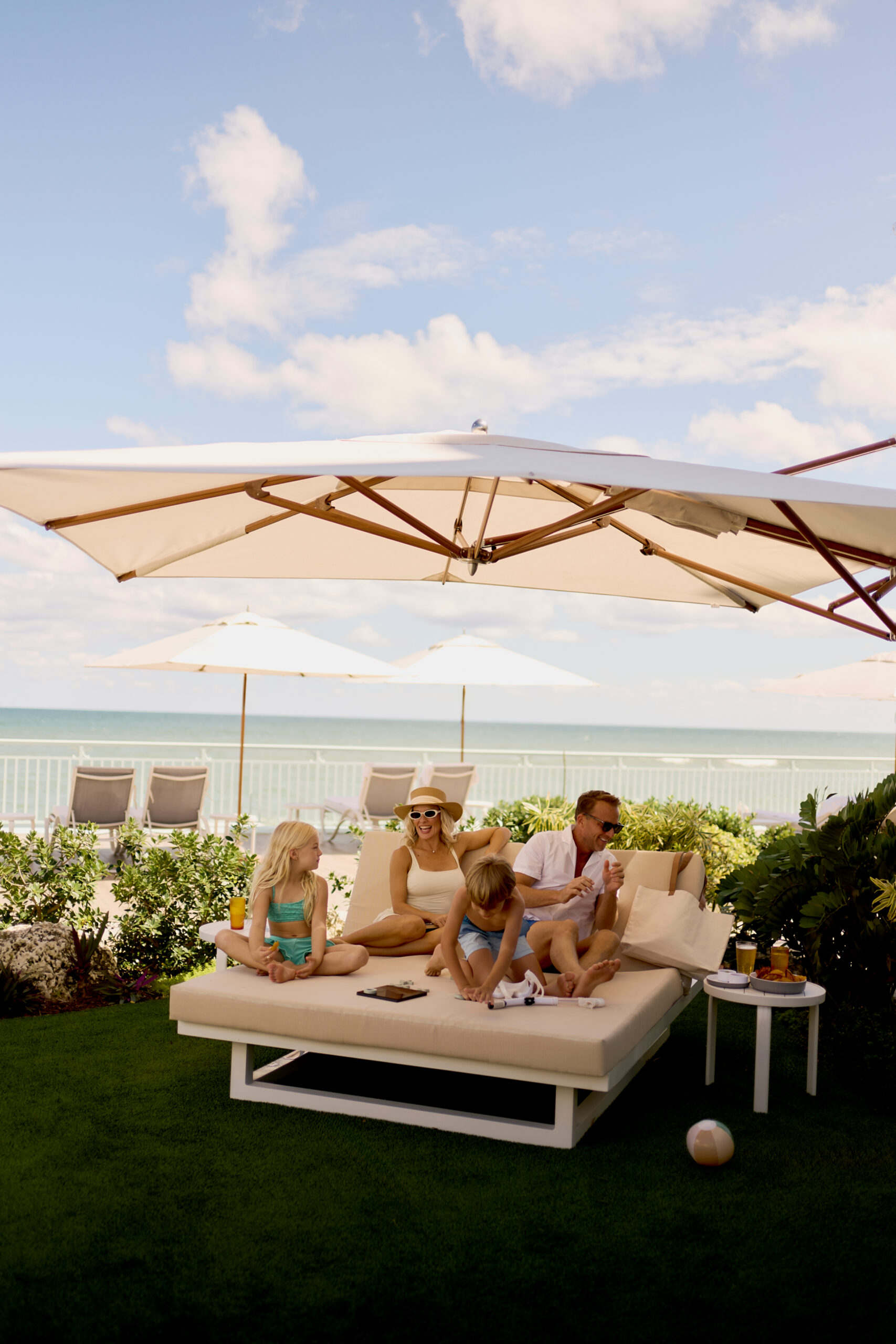 Family relaxing on outdoor daybed under umbrellas with ocean view in background