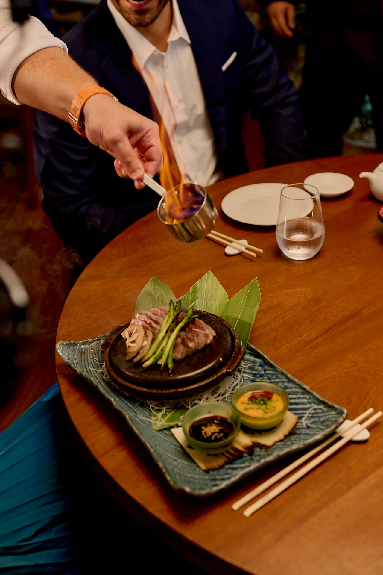 Dramatic tableside pour igniting a sizzling gourmet dish on a hot stone, with sauces and garnishes set for a refined dining experience