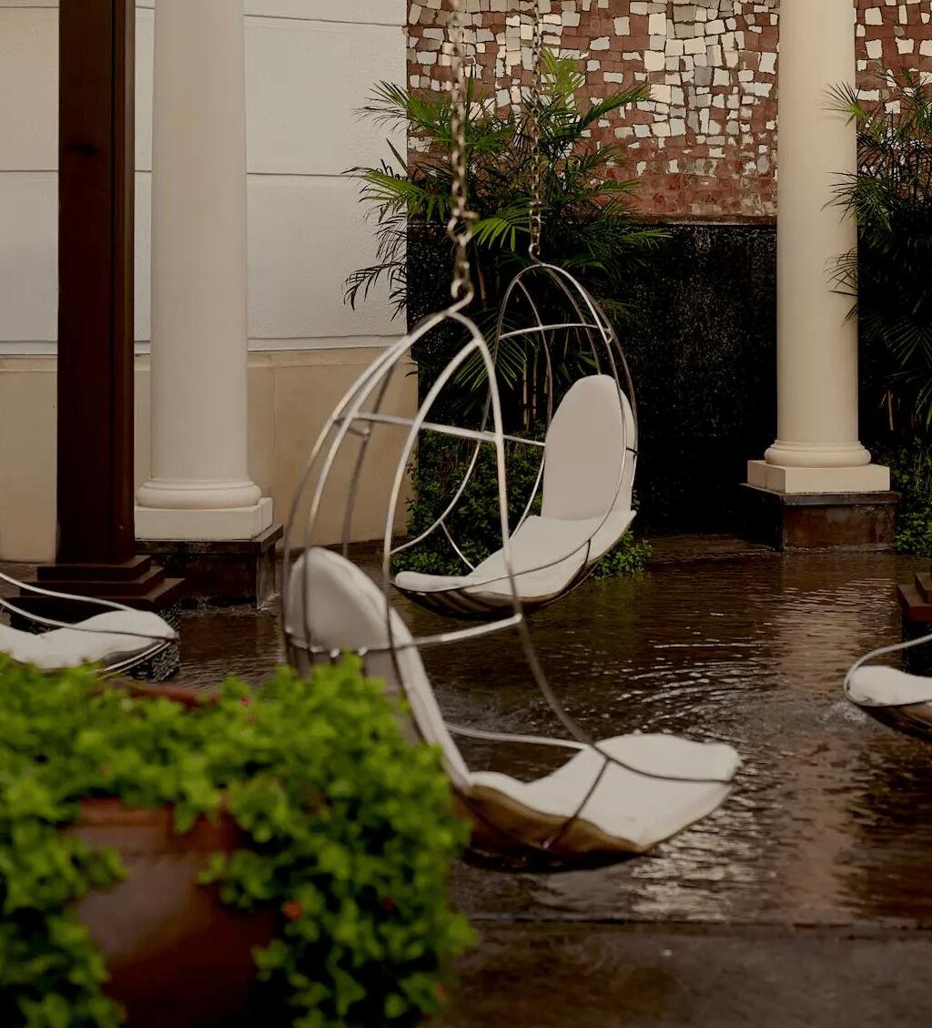 Outdoor lounge with modern hanging chairs under a wooden pergola, surrounded by columns and lush greenery