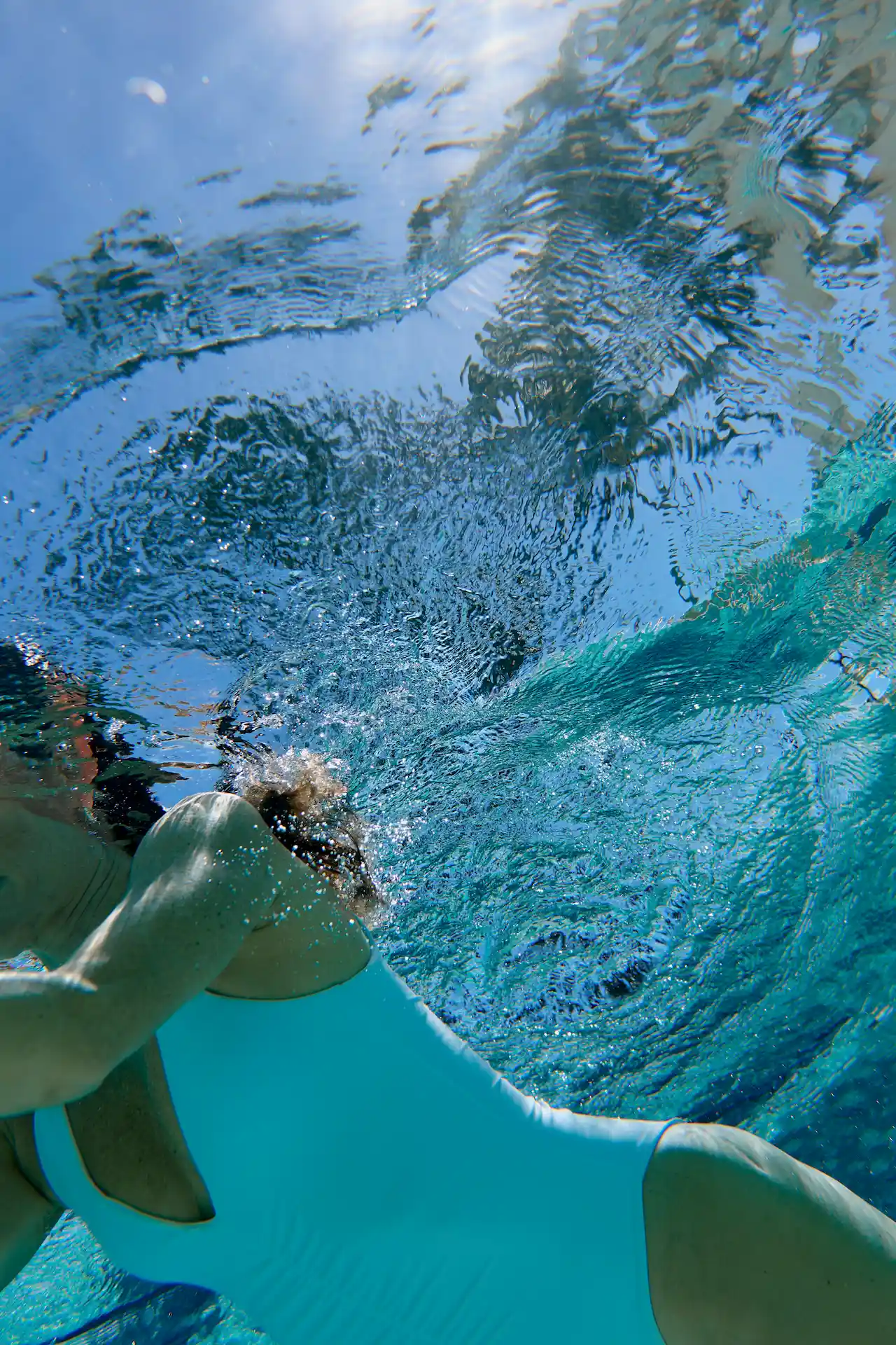 Underwater view of a swimmer in a light blue suit gliding beneath the surface as sunlight ripples above