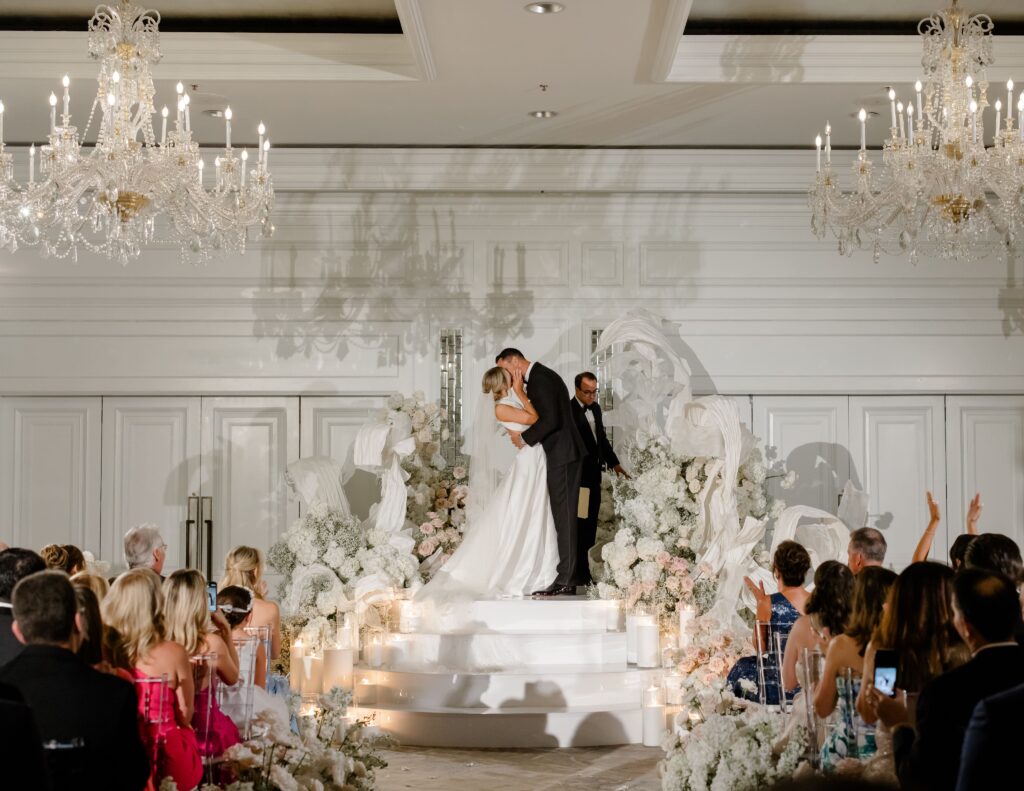 Bride and groom kissing on floral stage under chandeliers at elegant indoor wedding