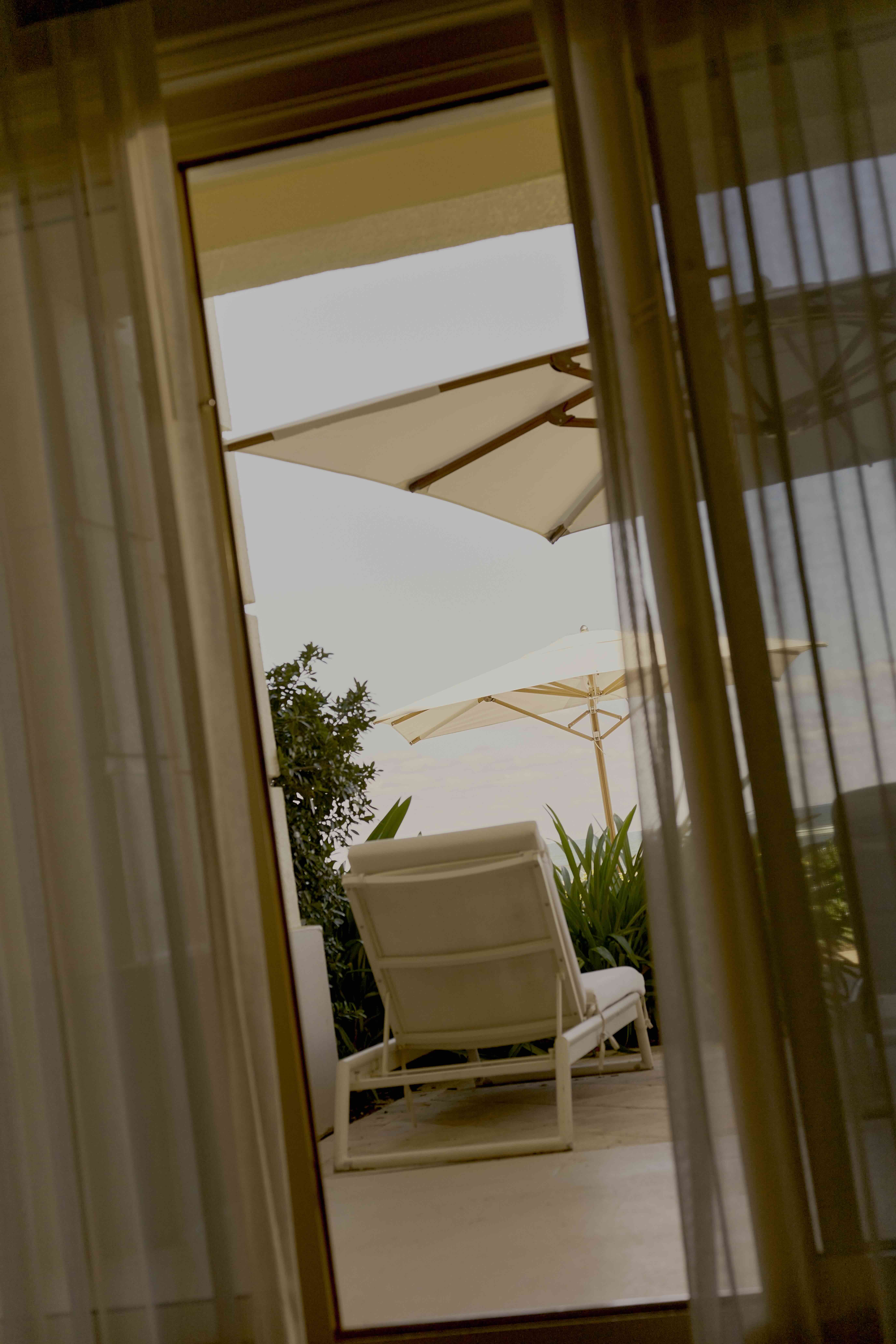 View through sheer curtains to patio lounge chair and white umbrellas overlooking the sea