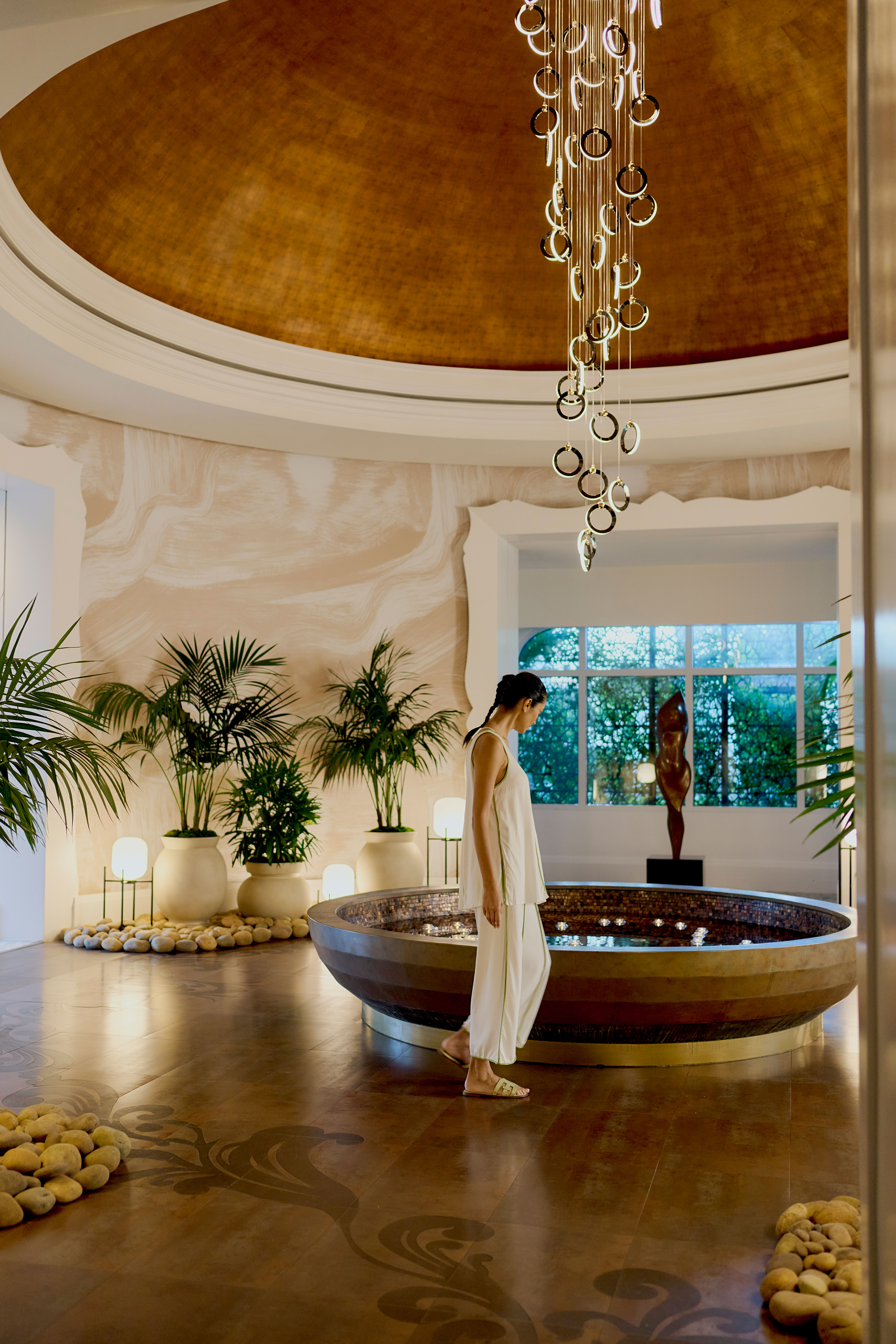 Woman walking beside circular indoor spa pool under golden dome ceiling with hanging chandelier