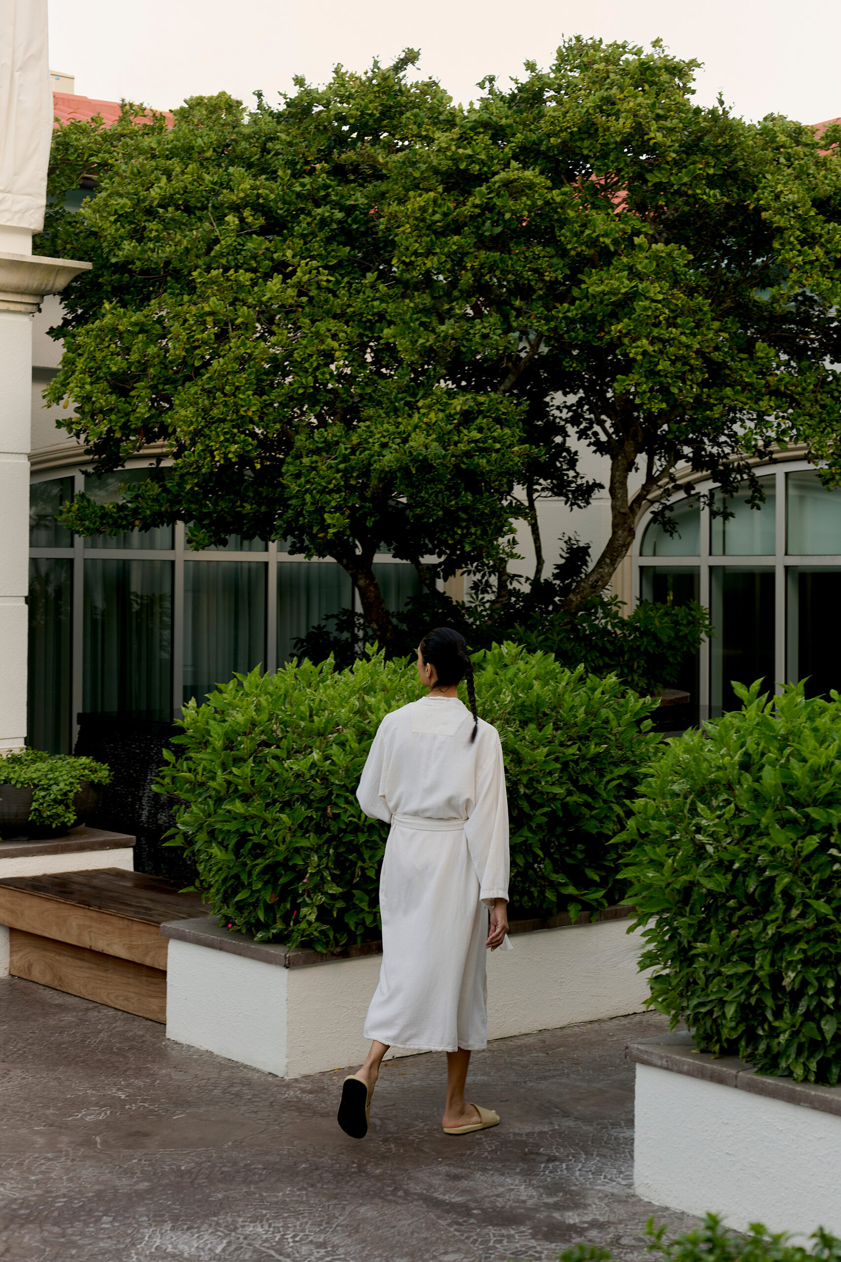 Woman in white robe walking through lush courtyard garden near modern building