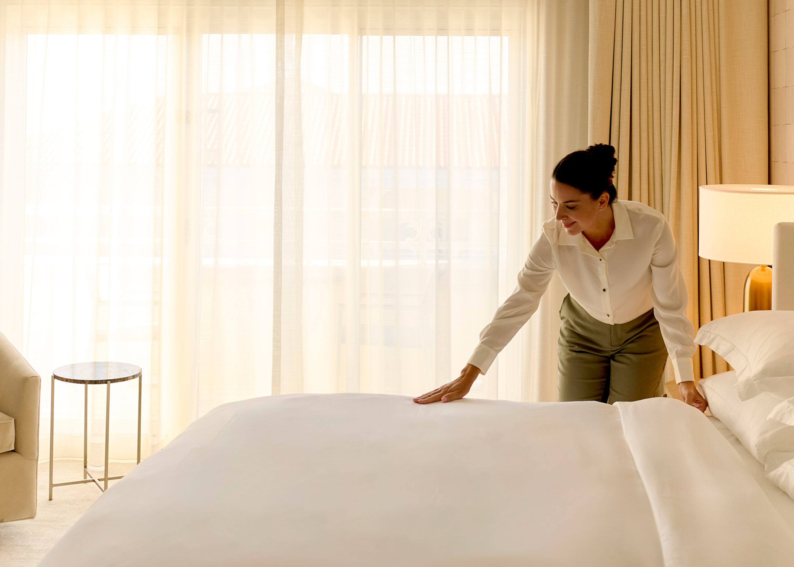Housekeeper smoothing white bedding in a sunlit hotel room with sheer curtains and bedside lamp