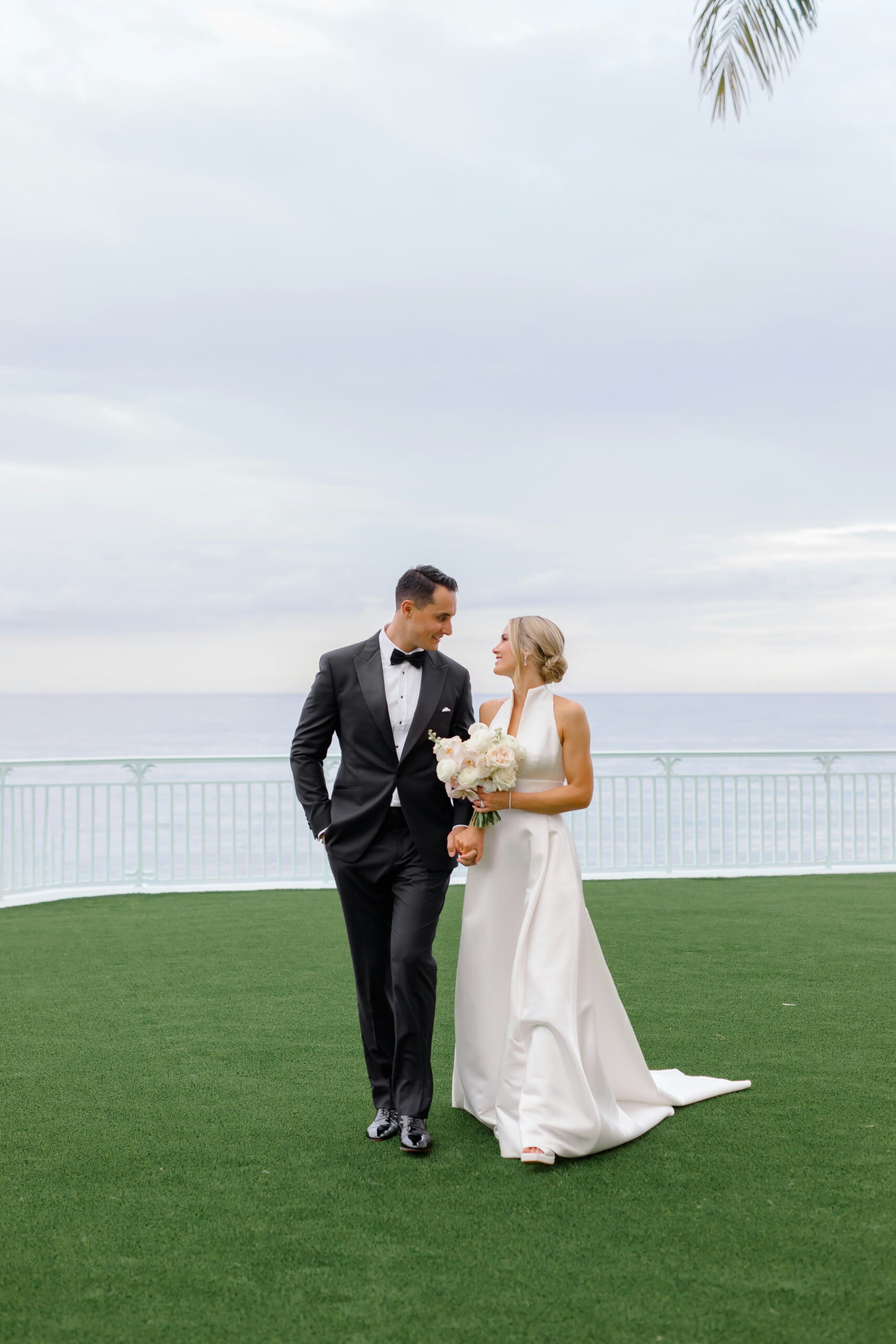 Bride and groom walking hand in hand on oceanfront lawn with bouquet